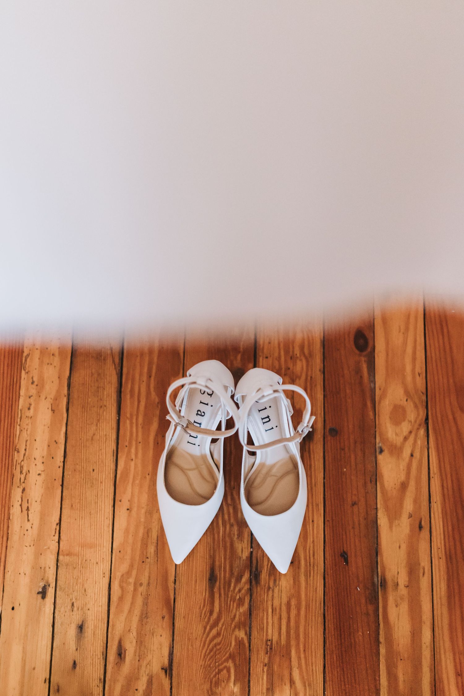 White pointed-toe ankle strap bridal heels on rustic hardwood floor.