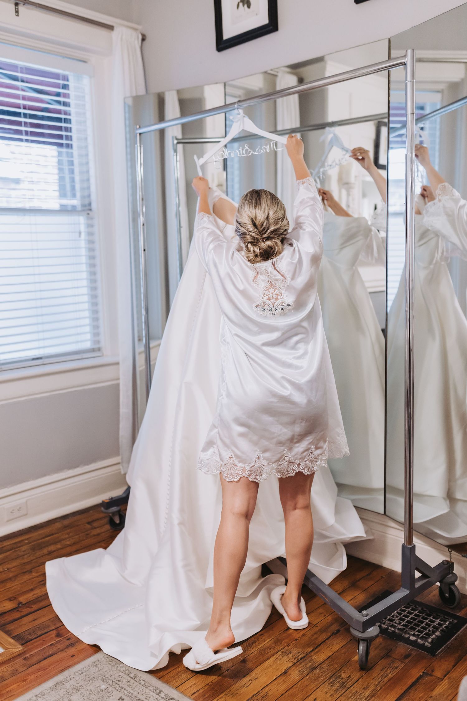 Bride in white satin robe reaching for her wedding gown on a garment rack before her ceremony.