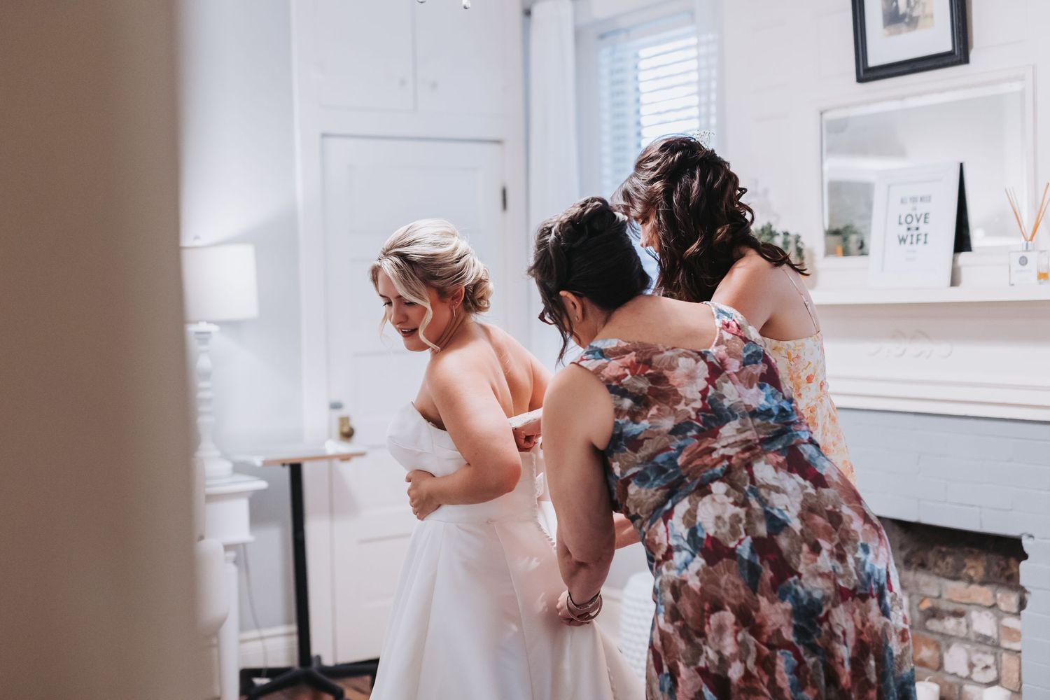 Two bridesmaids help a bride button up her white wedding gown in a bright elegant bridal suite.