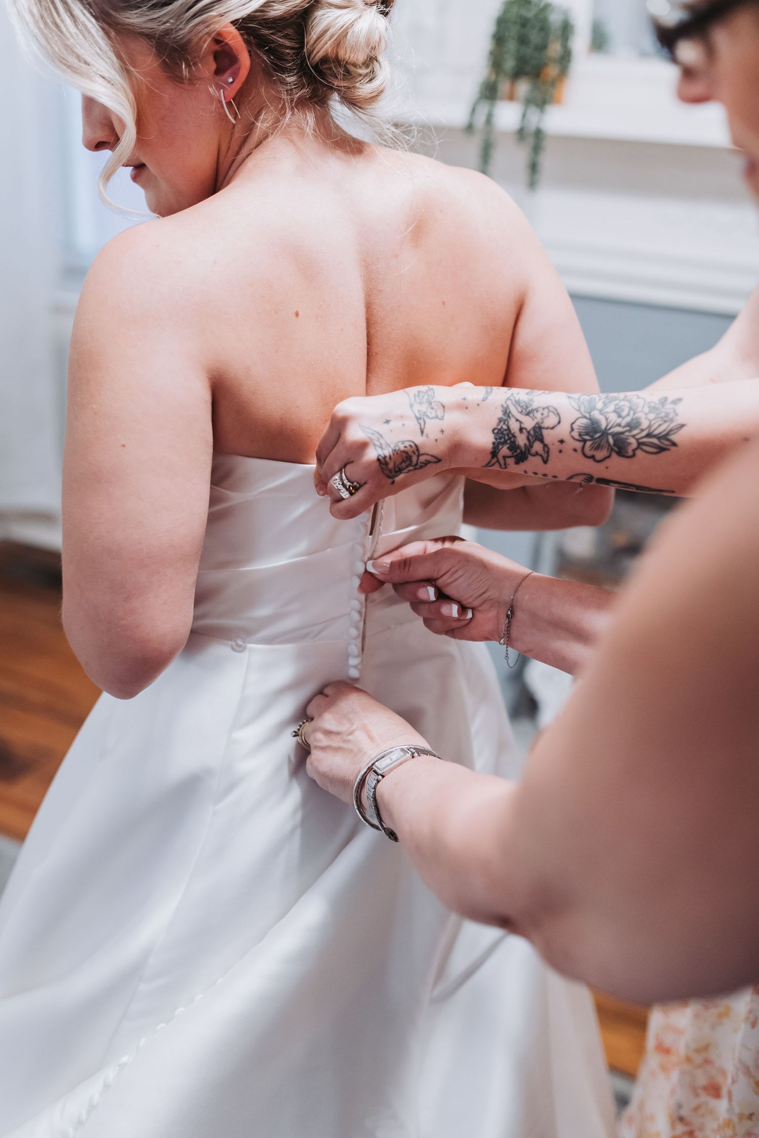 Close-up of tattooed hands carefully fastening buttons on the back of a white strapless wedding dress.