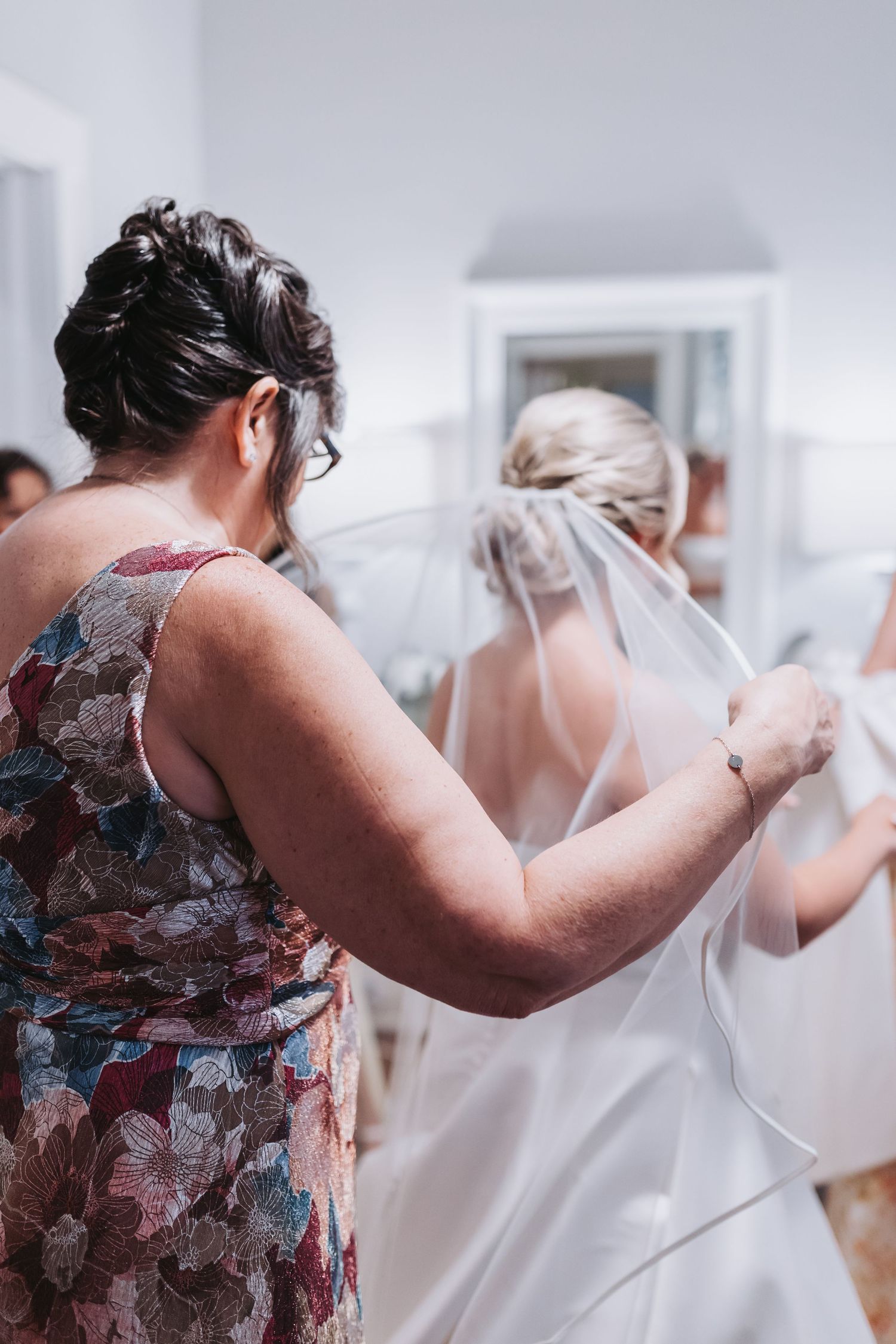 Woman in floral dress helping bride with wedding veil during bridal preparations.