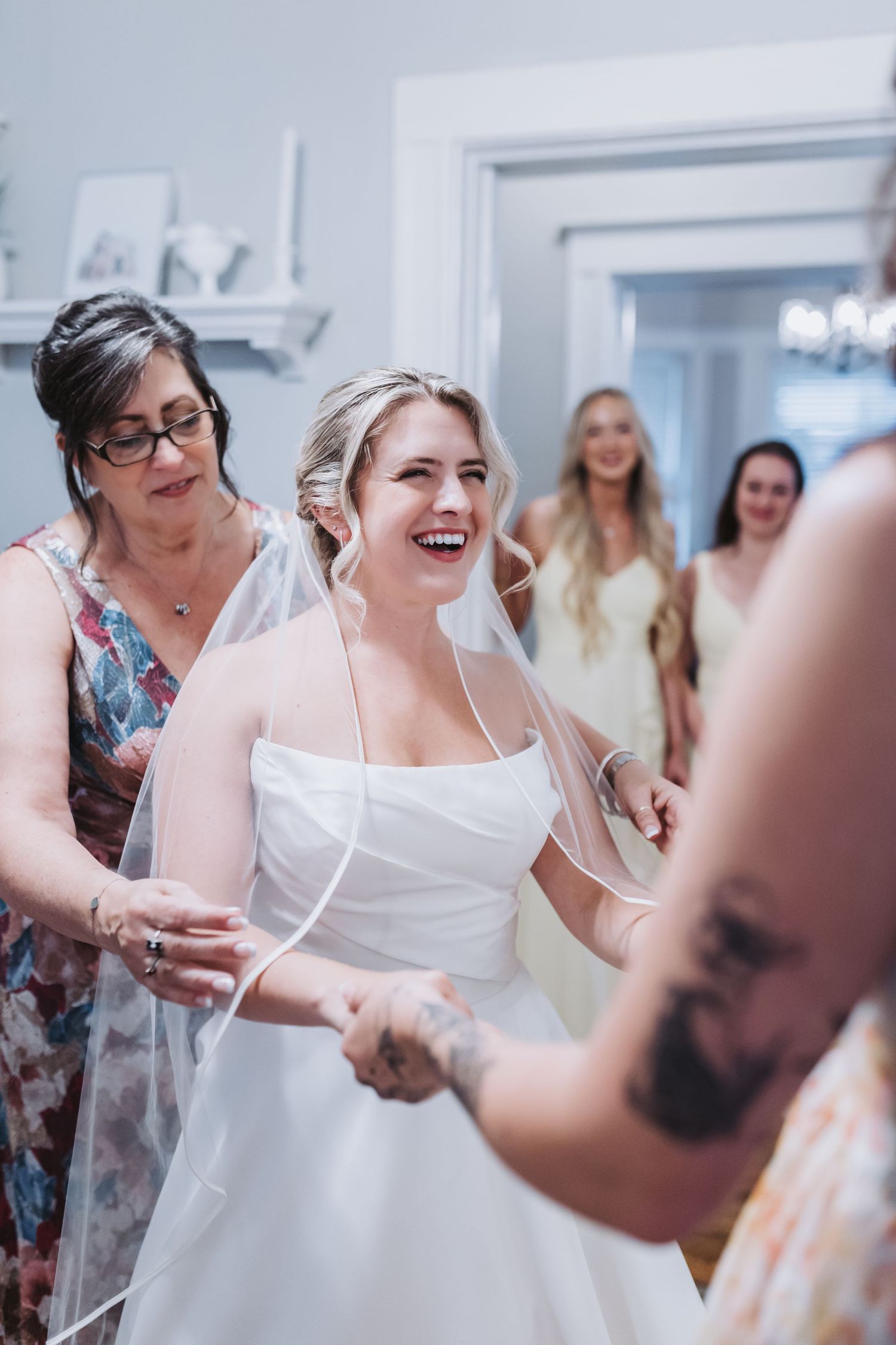 Joyful bride in white gown laughing with family and bridesmaids during wedding prep.