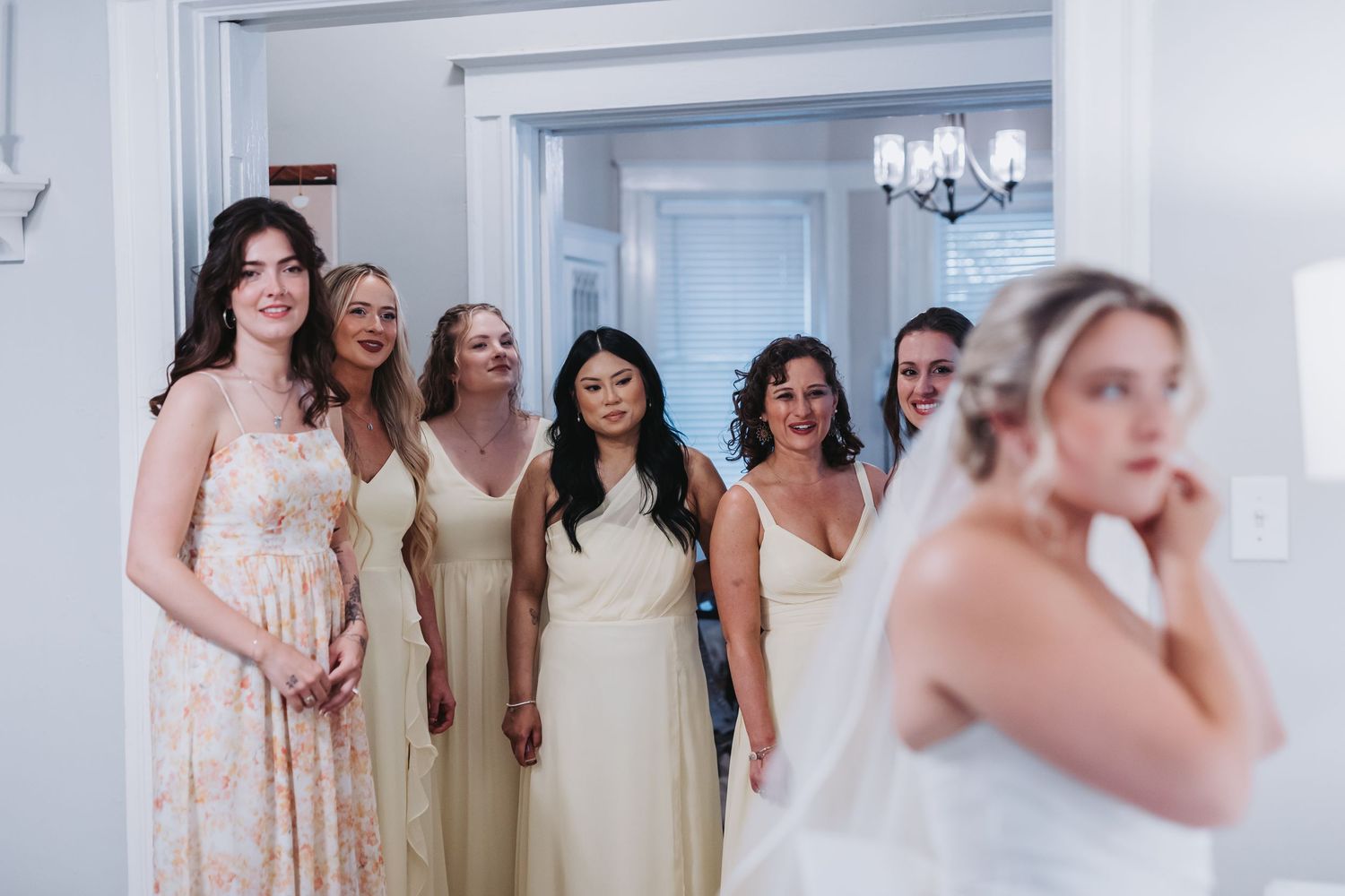 Bridesmaids in yellow dresses reacting with joy as bride in white gown and veil stands nearby.