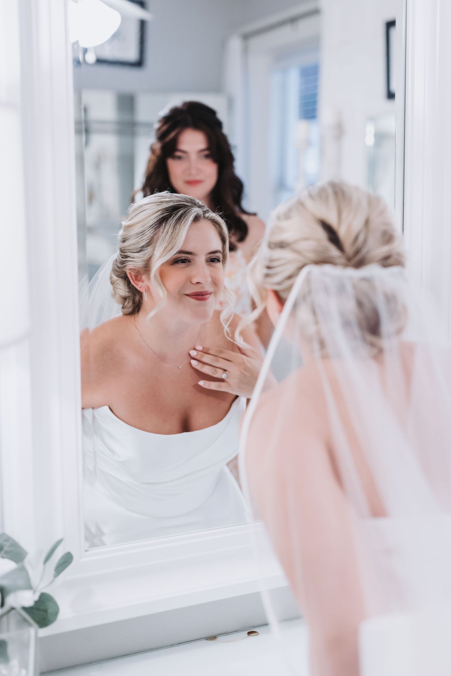 Blonde bride in white gown admires her reflection in a mirror while a bridesmaid looks on behind her.