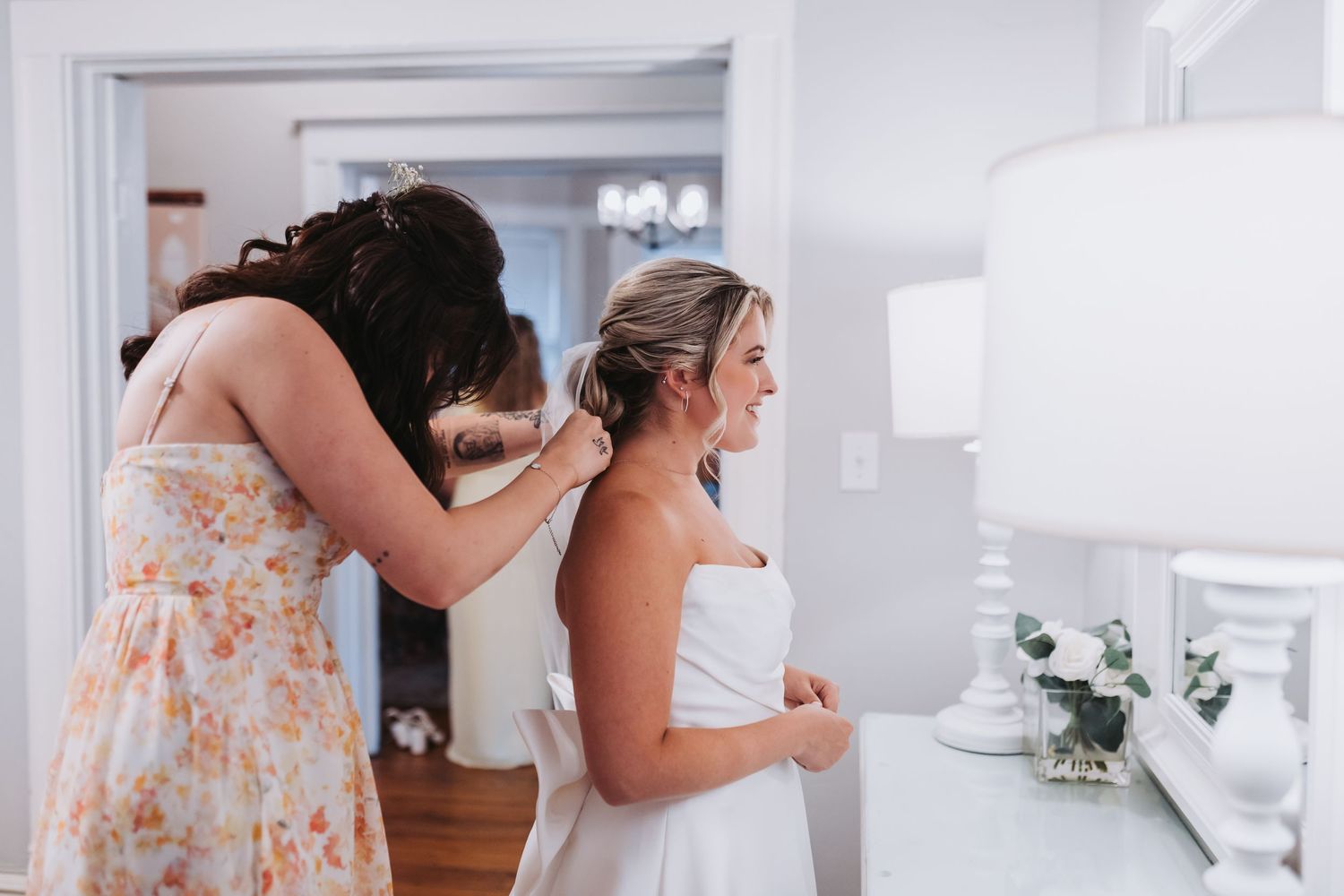Bridesmaid in floral dress helping bride with her white strapless wedding gown in a bright dressing room.