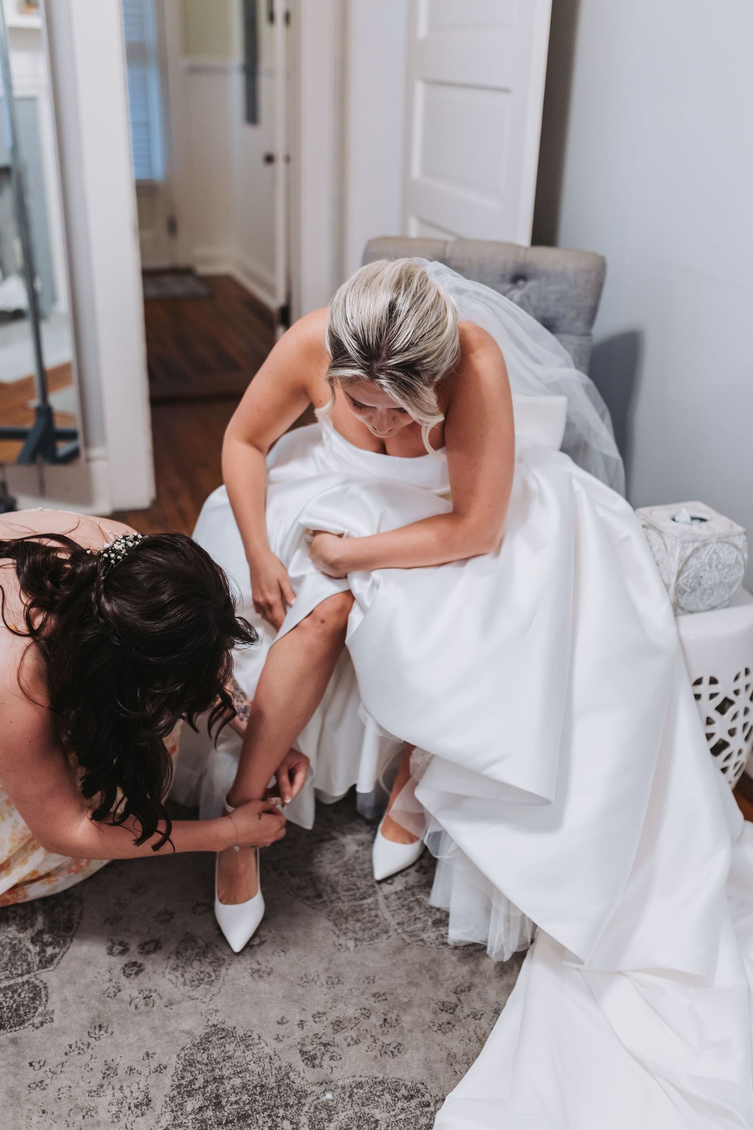 Bridesmaid helping bride put on white heels while bride sits in wedding dress before the ceremony.