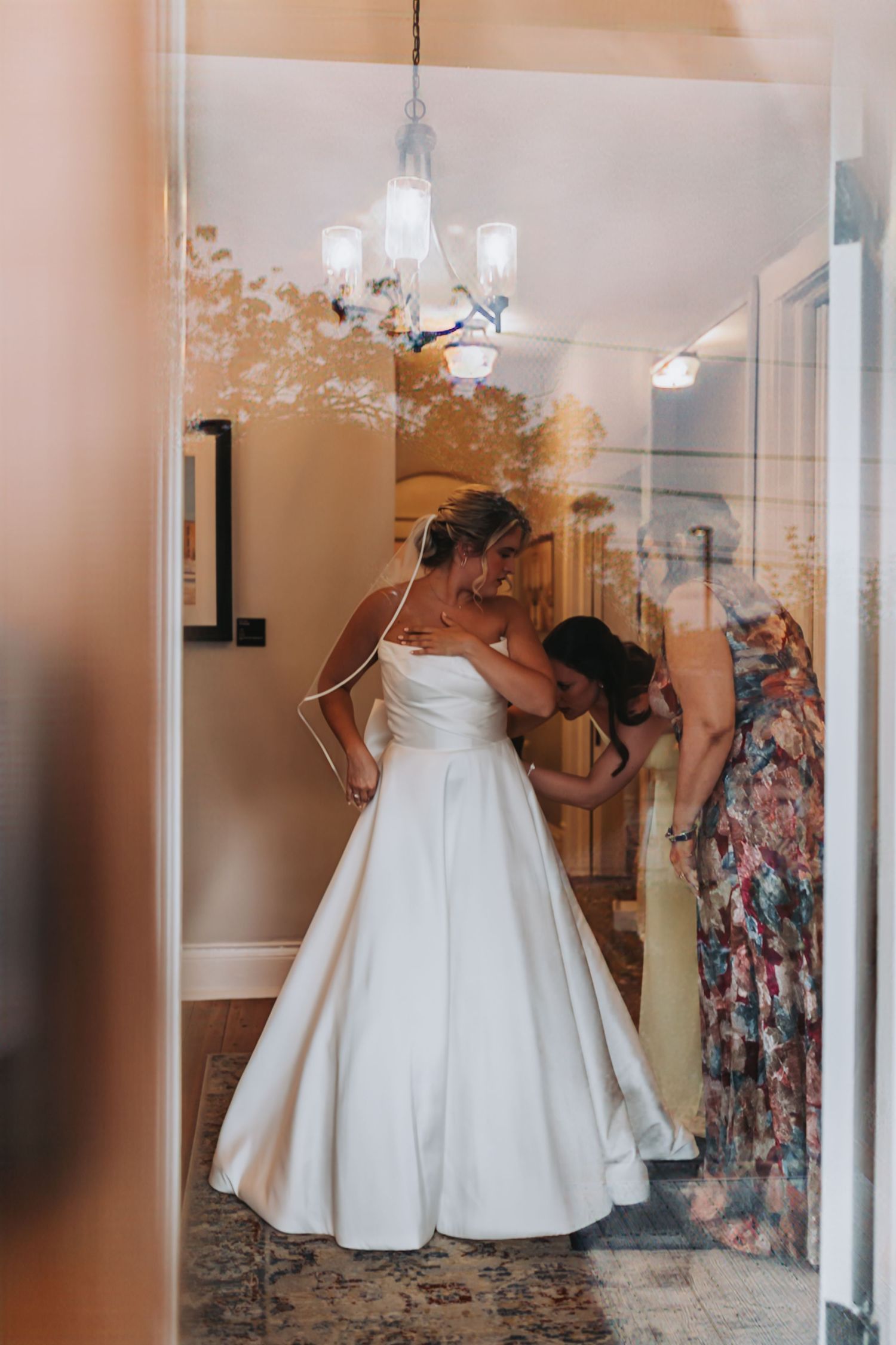 Bride in white wedding gown being helped by bridesmaids in yellow and floral dresses inside an elegant room.