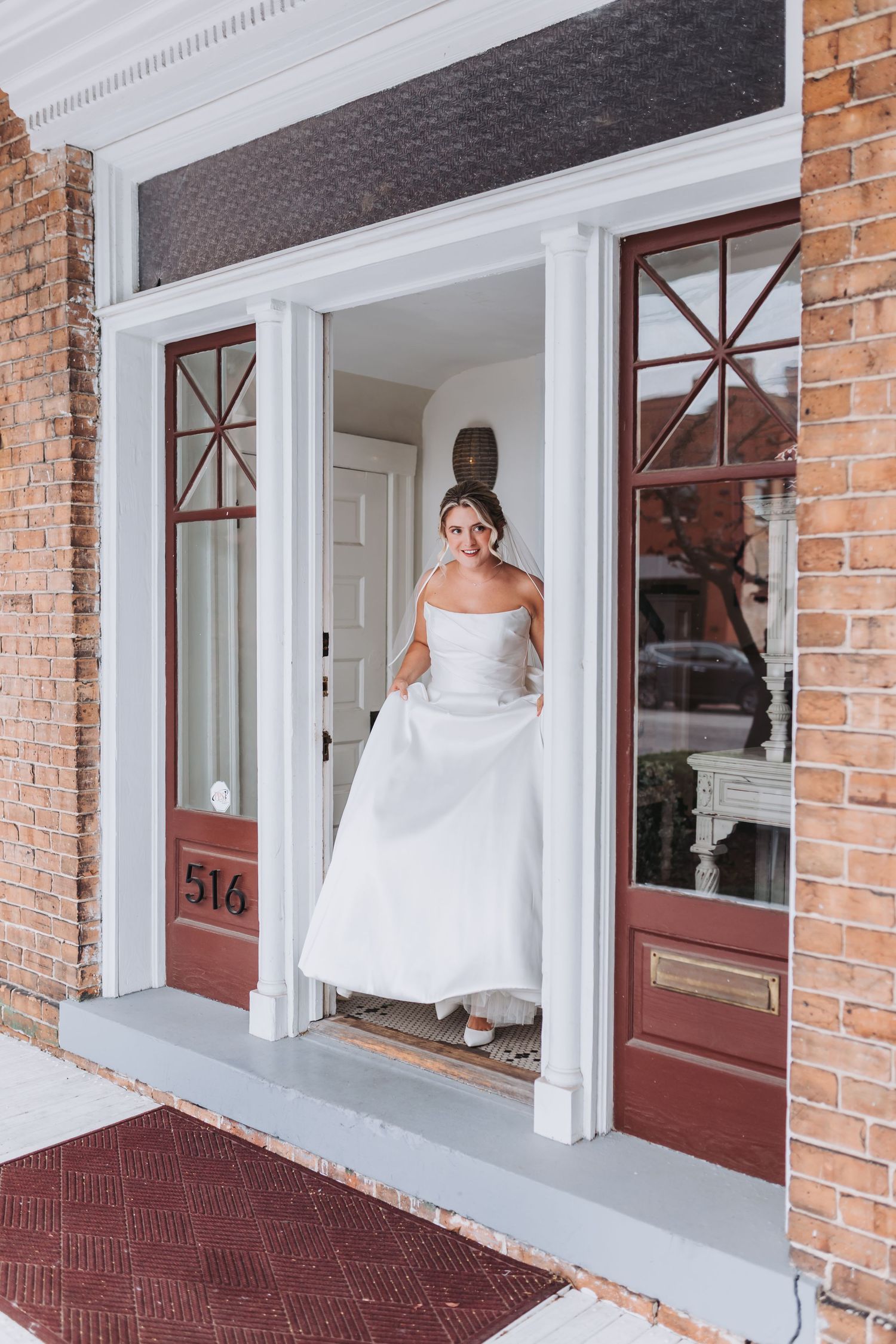 Smiling bride in a white strapless ball gown stands in the doorway of a classic brick building numbered 514.