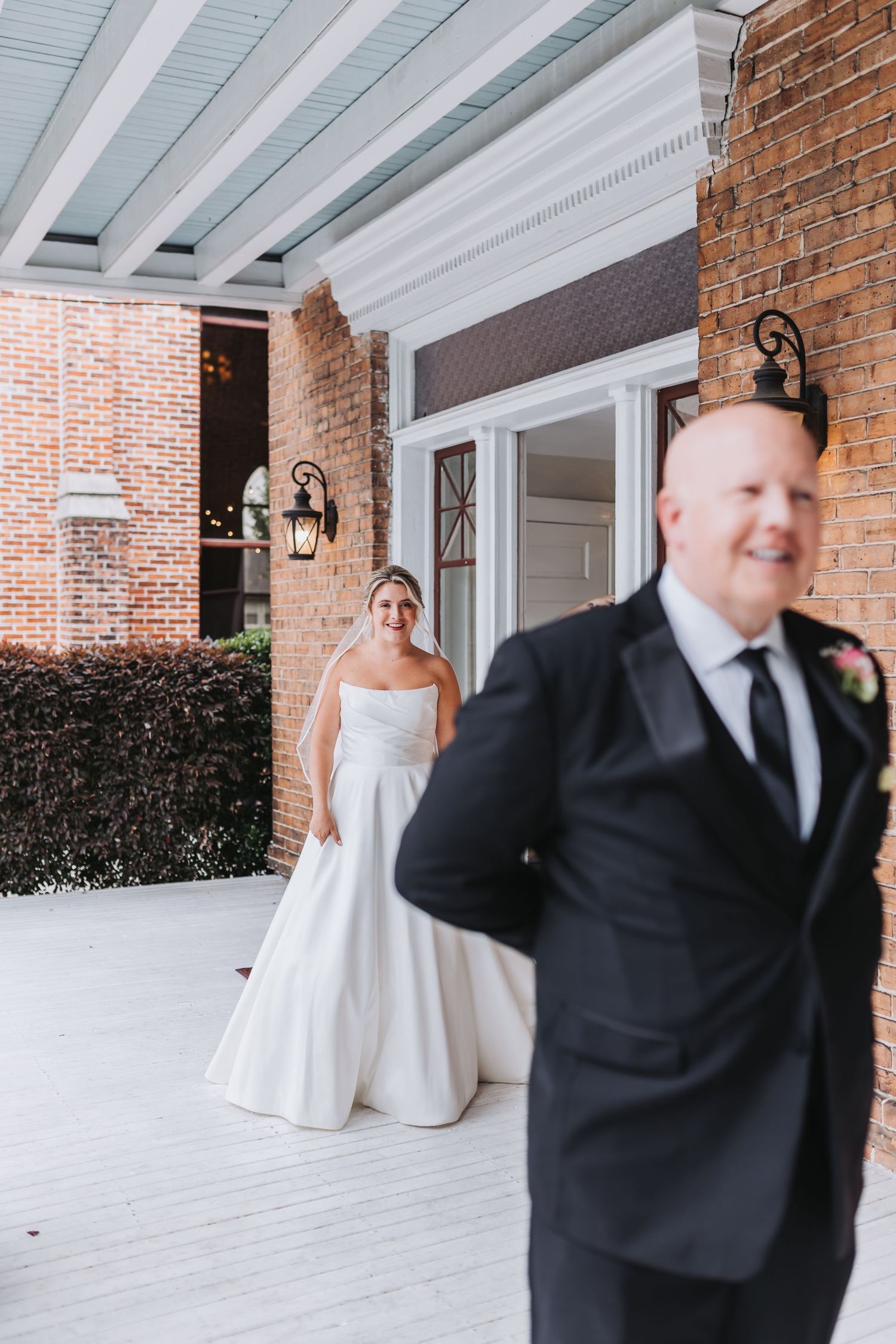 Bride in white strapless gown smiles at groom in black tuxedo during wedding first look on brick building porch.