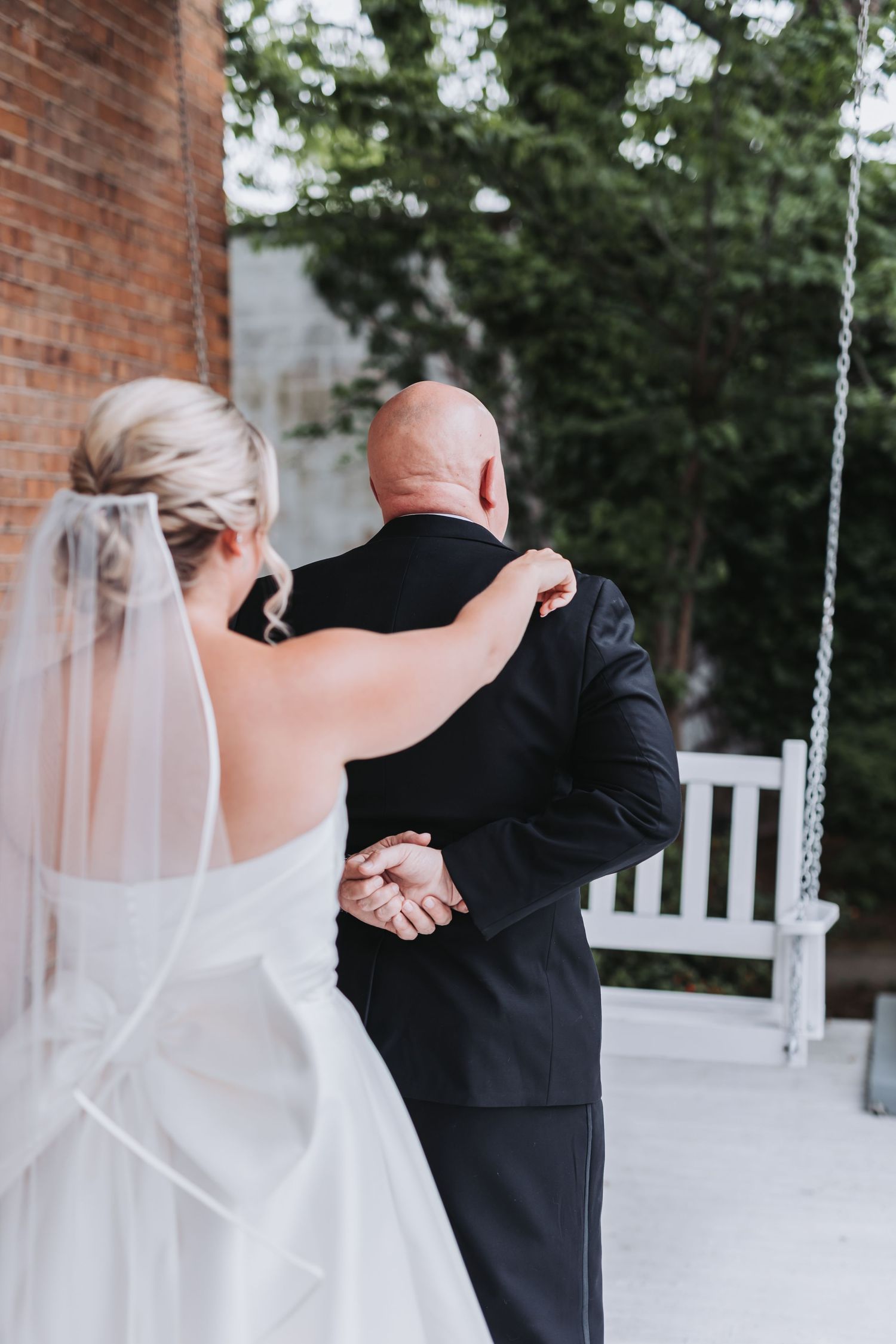 Bride in white gown and veil holds hands with bald groom in black suit during outdoor first look.