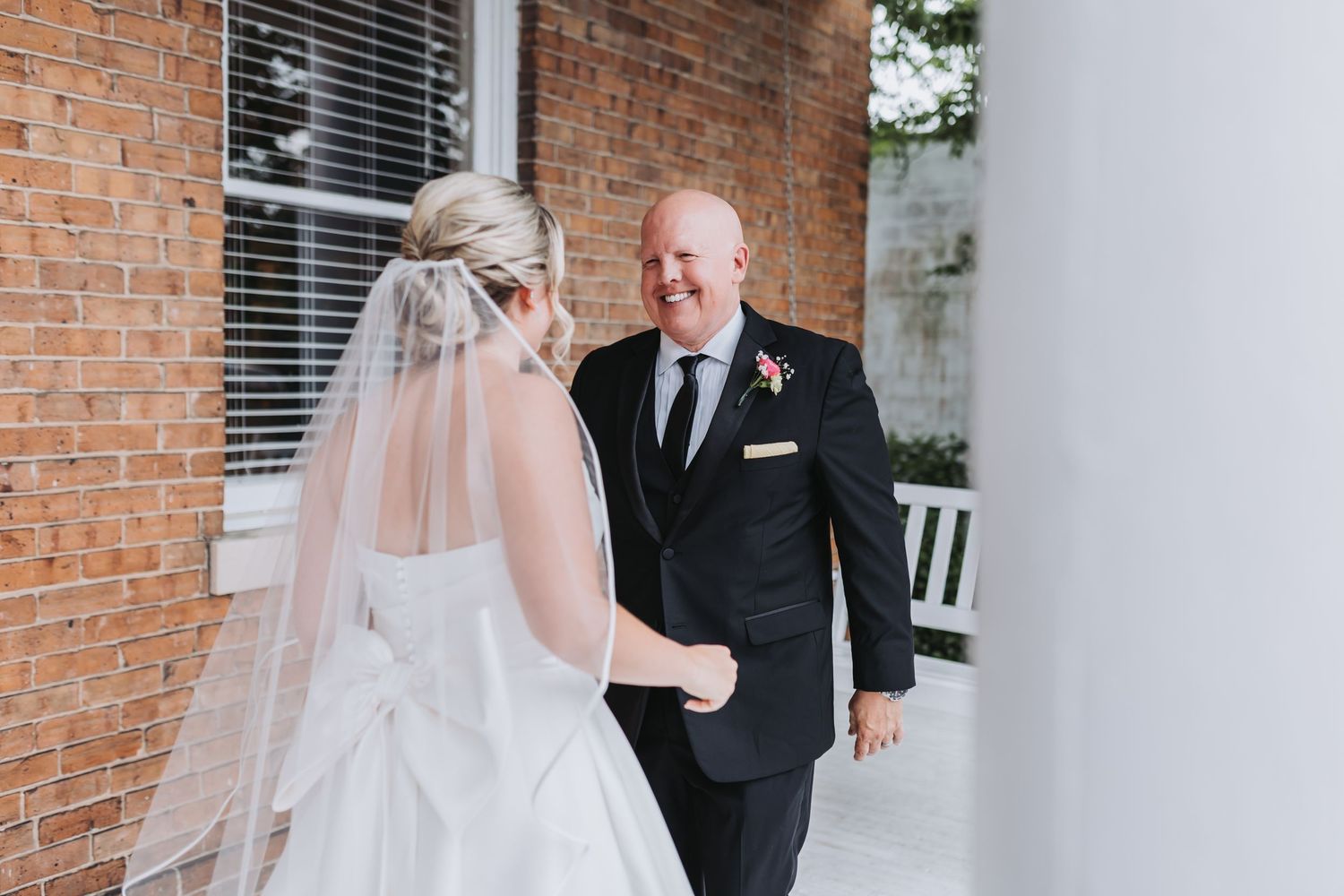 Emotional father-daughter first look moment outside a brick building as bride approaches in white gown and veil.