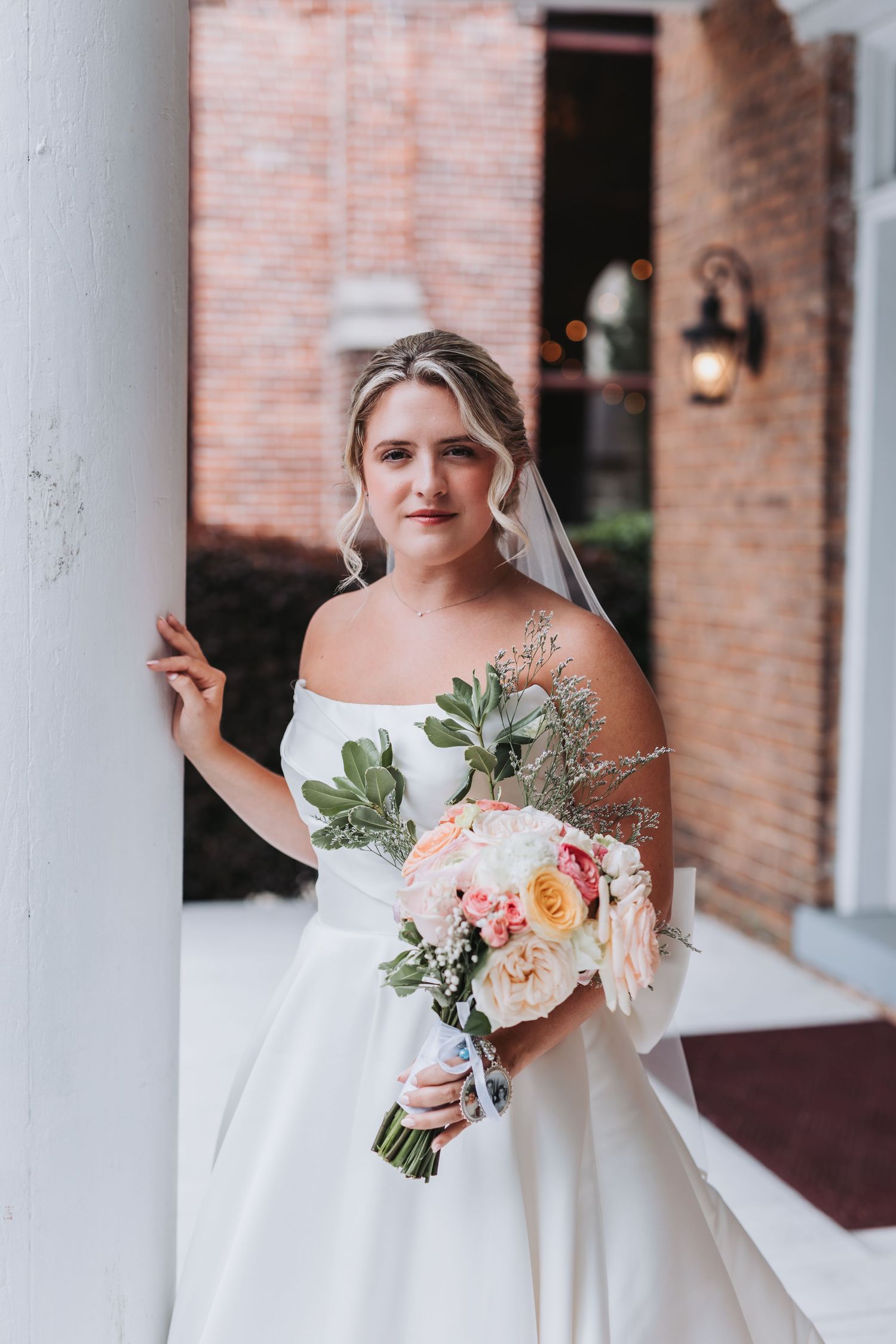 Bride in strapless white gown holding pink and peach bouquet leaning against white column.