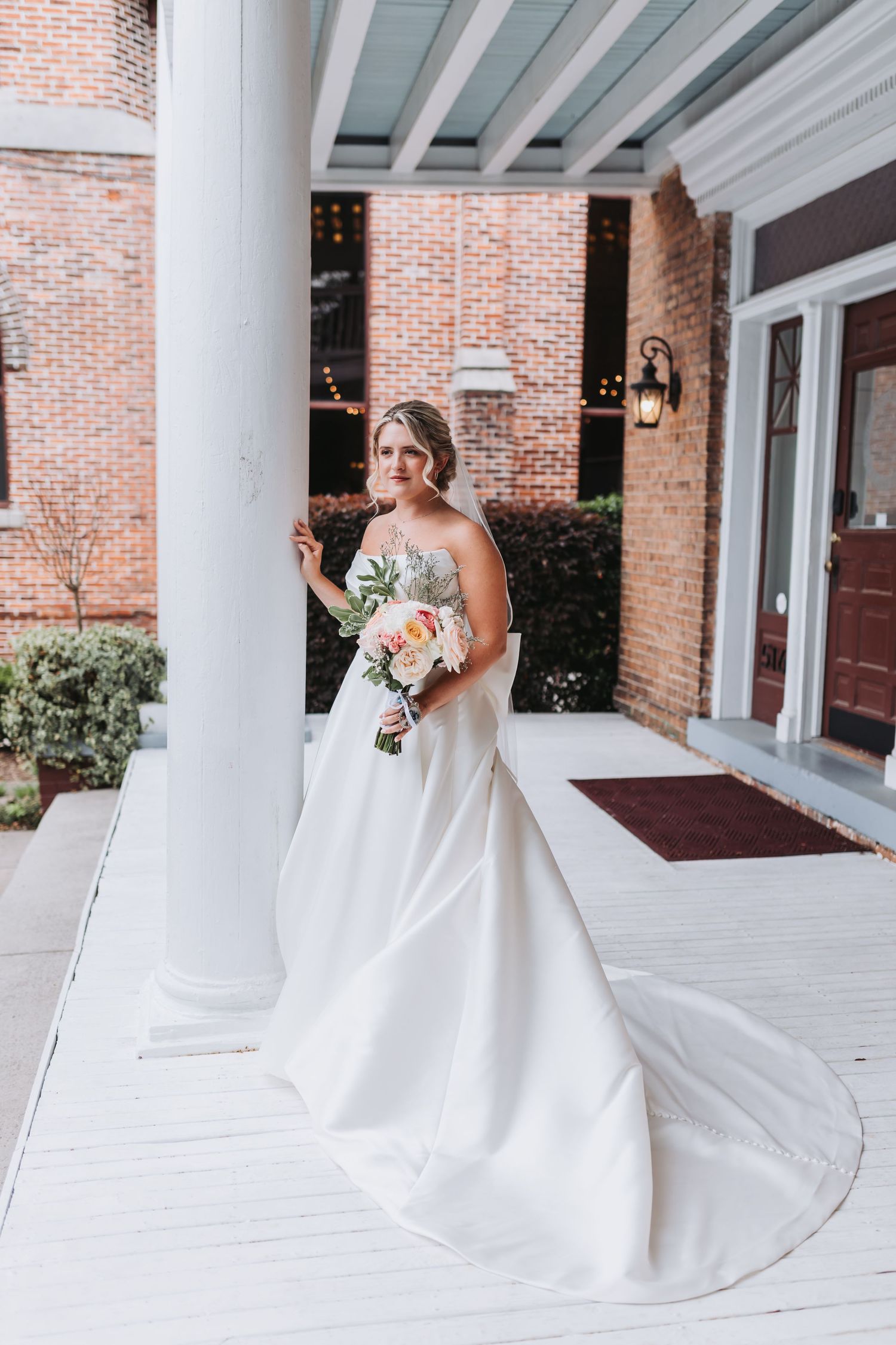 Elegant bride in white strapless gown holding pink bouquet on venue porch.