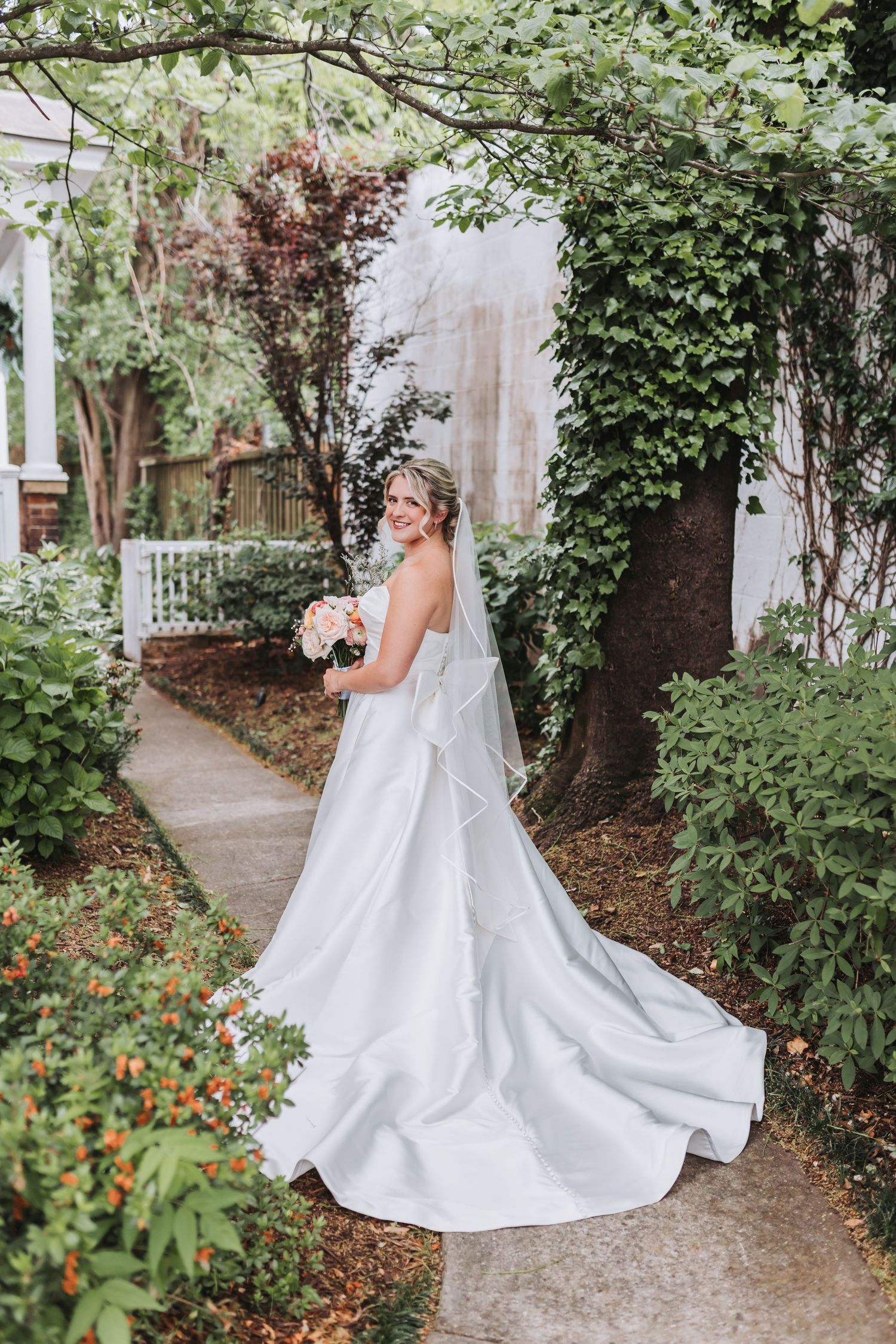 Bride in white gown with long train holds bouquet on garden path.