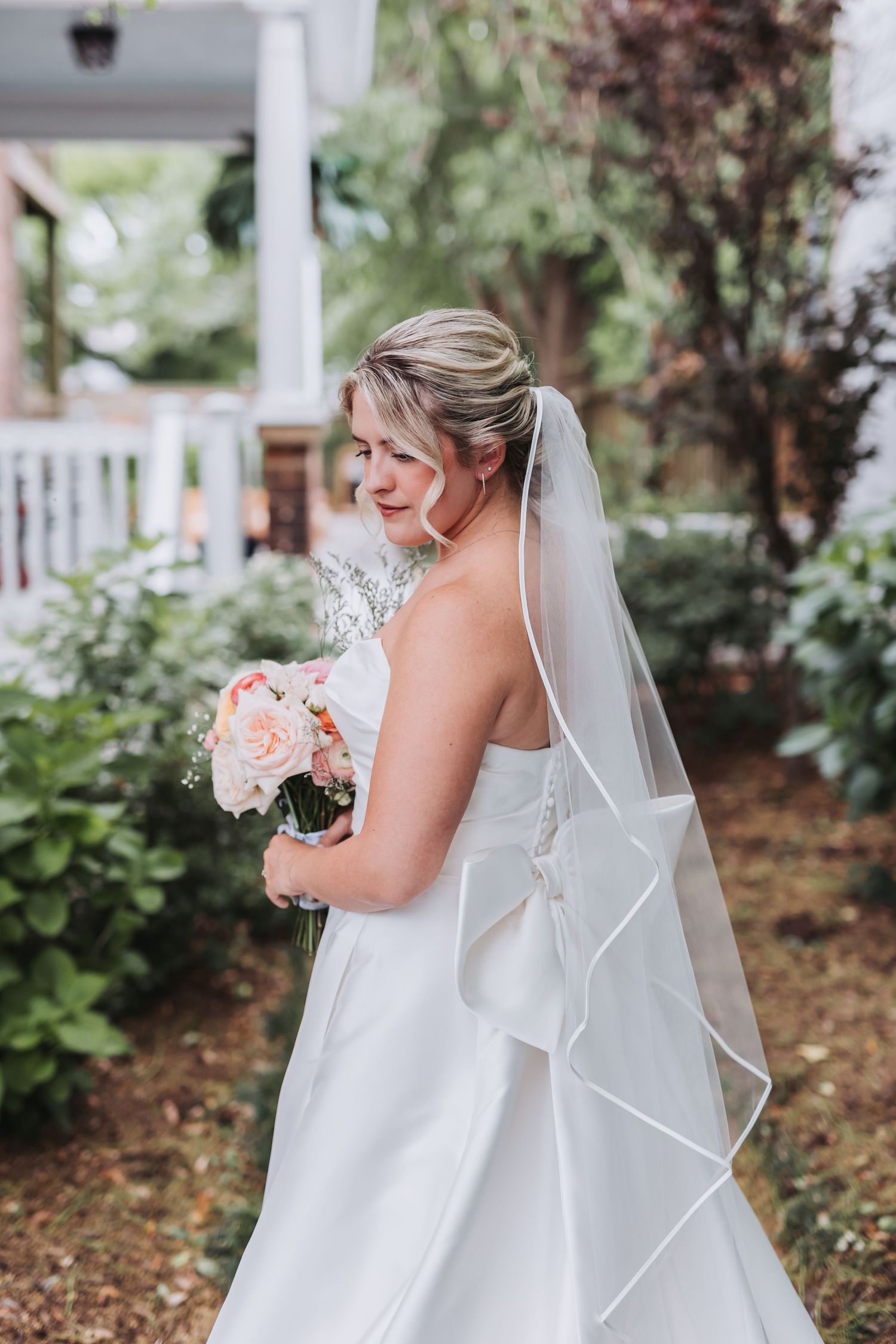 Bride in white gown with bow and veil holds blush bouquet, smiling over shoulder in lush garden setting.