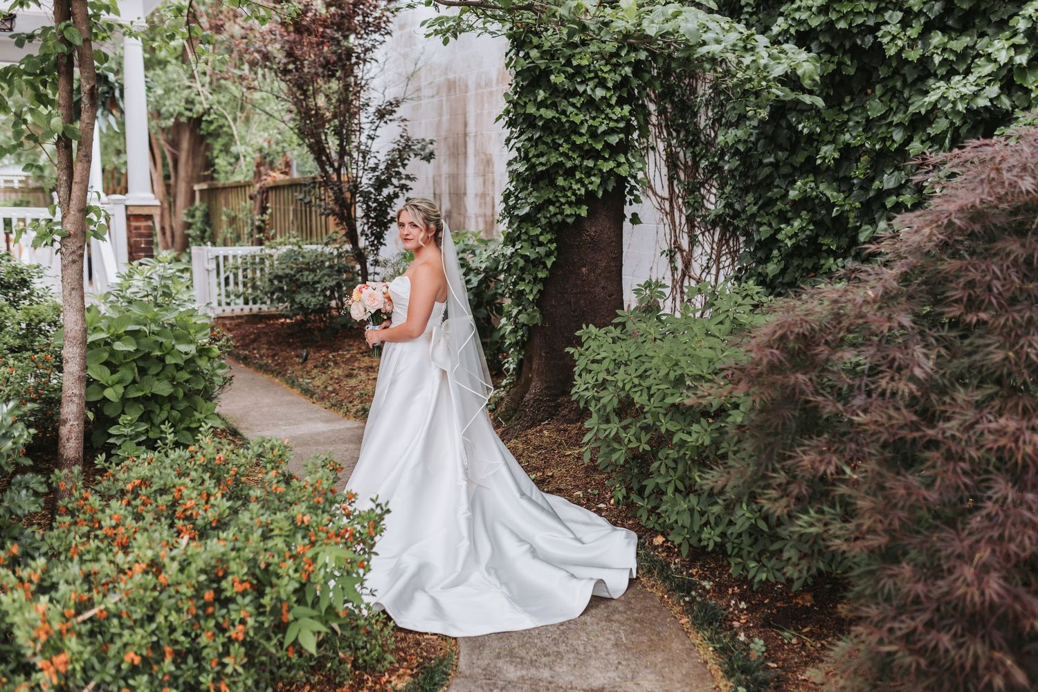 Bride in a flowing white ball gown and veil poses in a lush garden holding a soft pink bouquet.