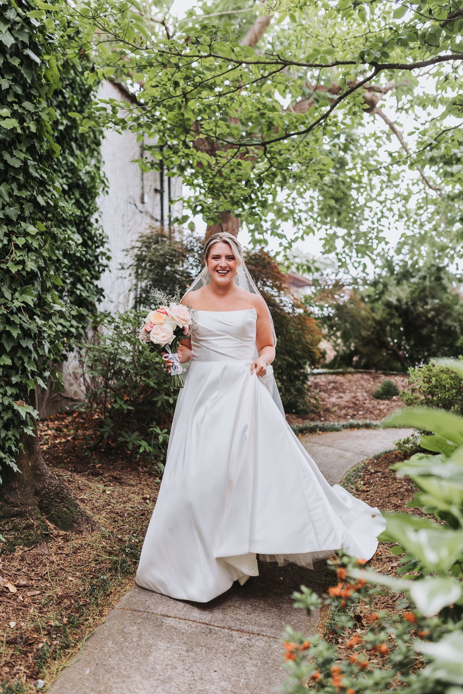 Radiant bride in a white strapless ball gown walks along a garden path holding a pink and peach bouquet.