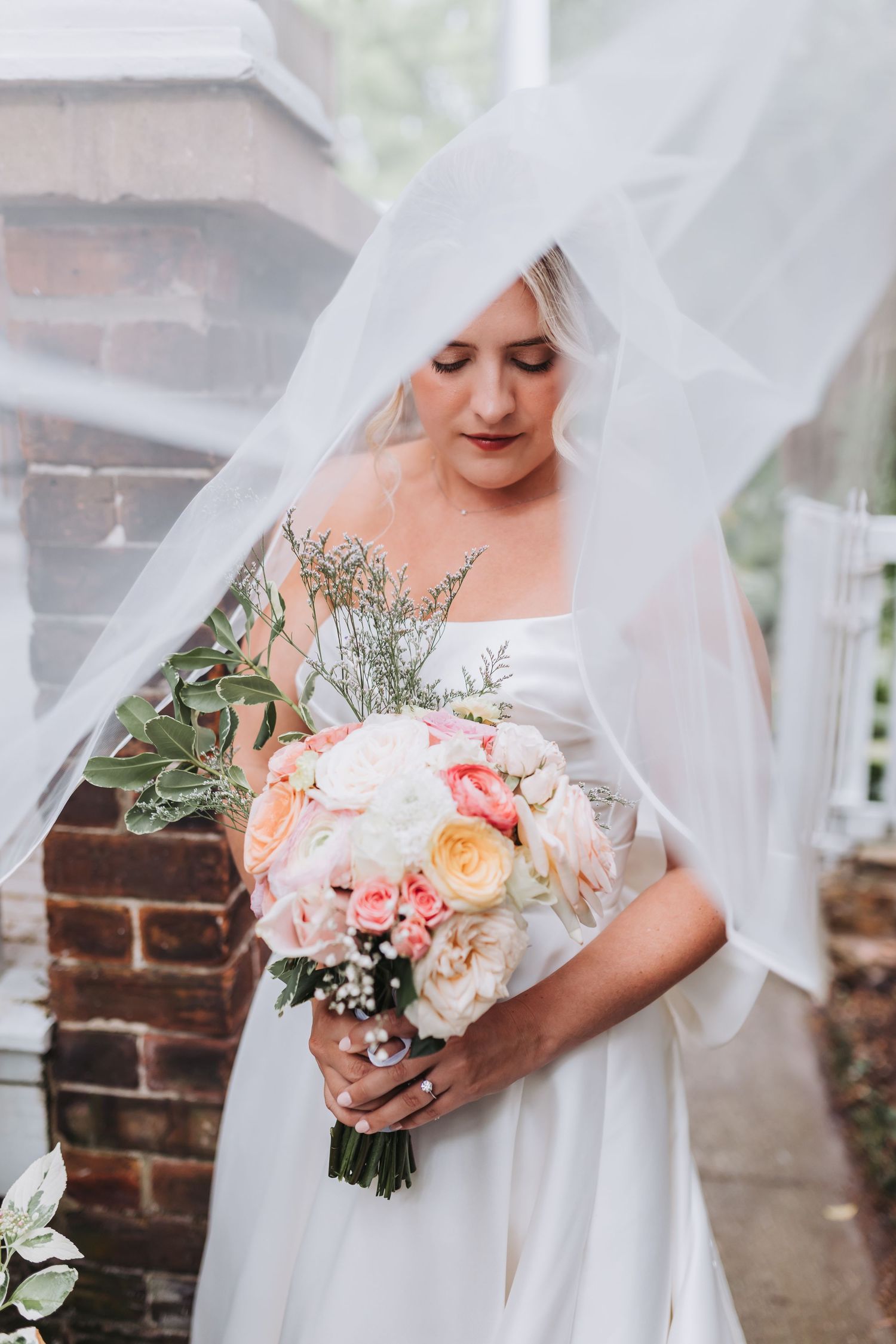 Bride in white gown holds pink and white bouquet under flowing veil near brick column.