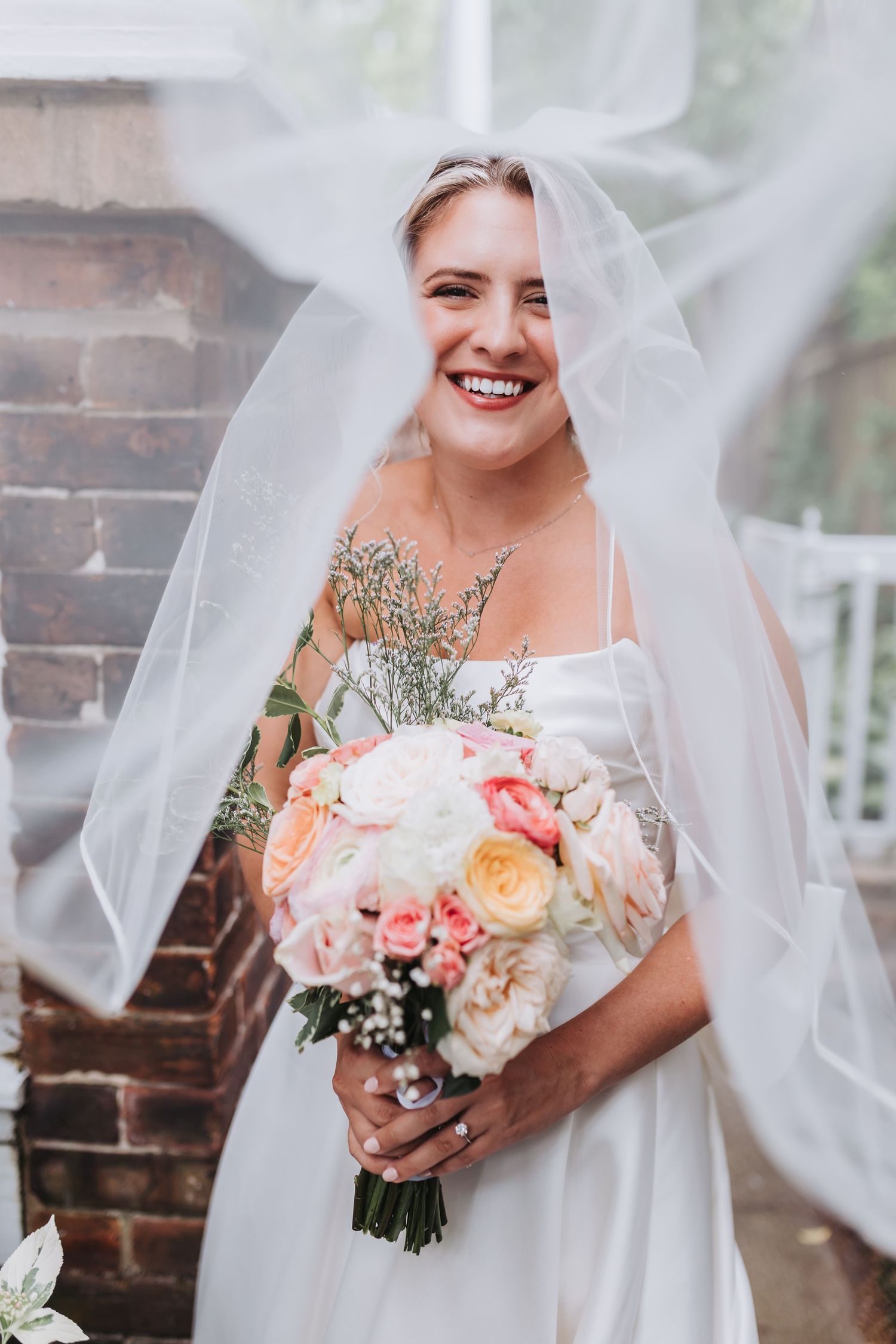 Joyful bride laughing with veil blowing in the wind while holding a pink and white floral bouquet.