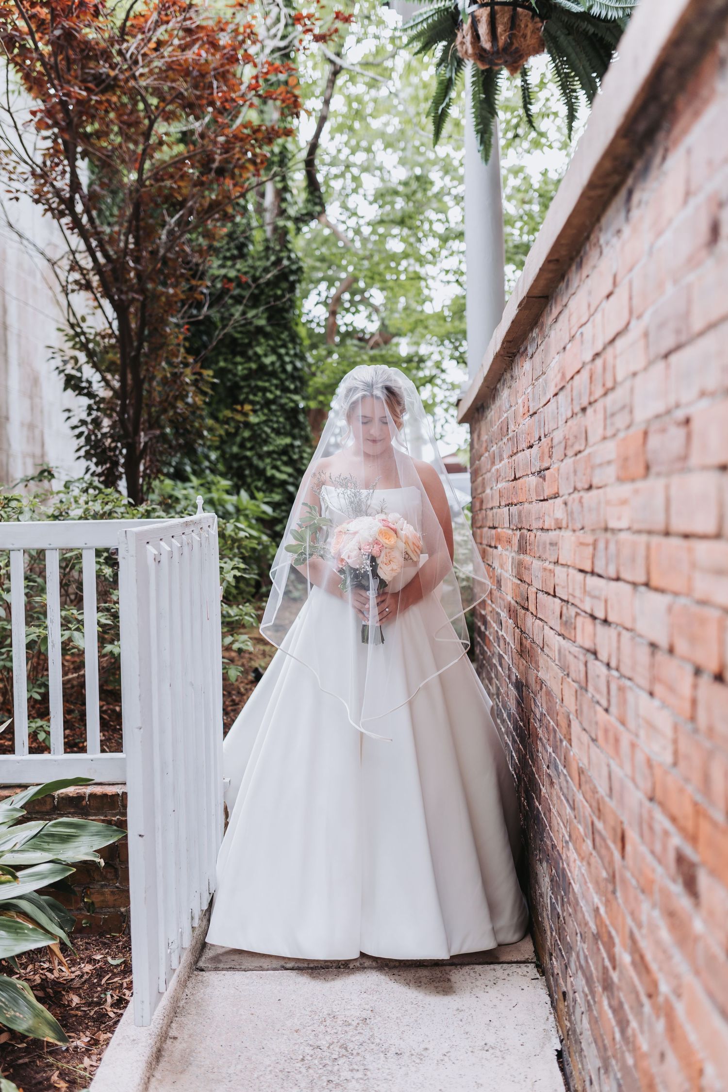 Elegant bride in a white ball gown and veil stands beside a rustic brick wall in a garden setting.