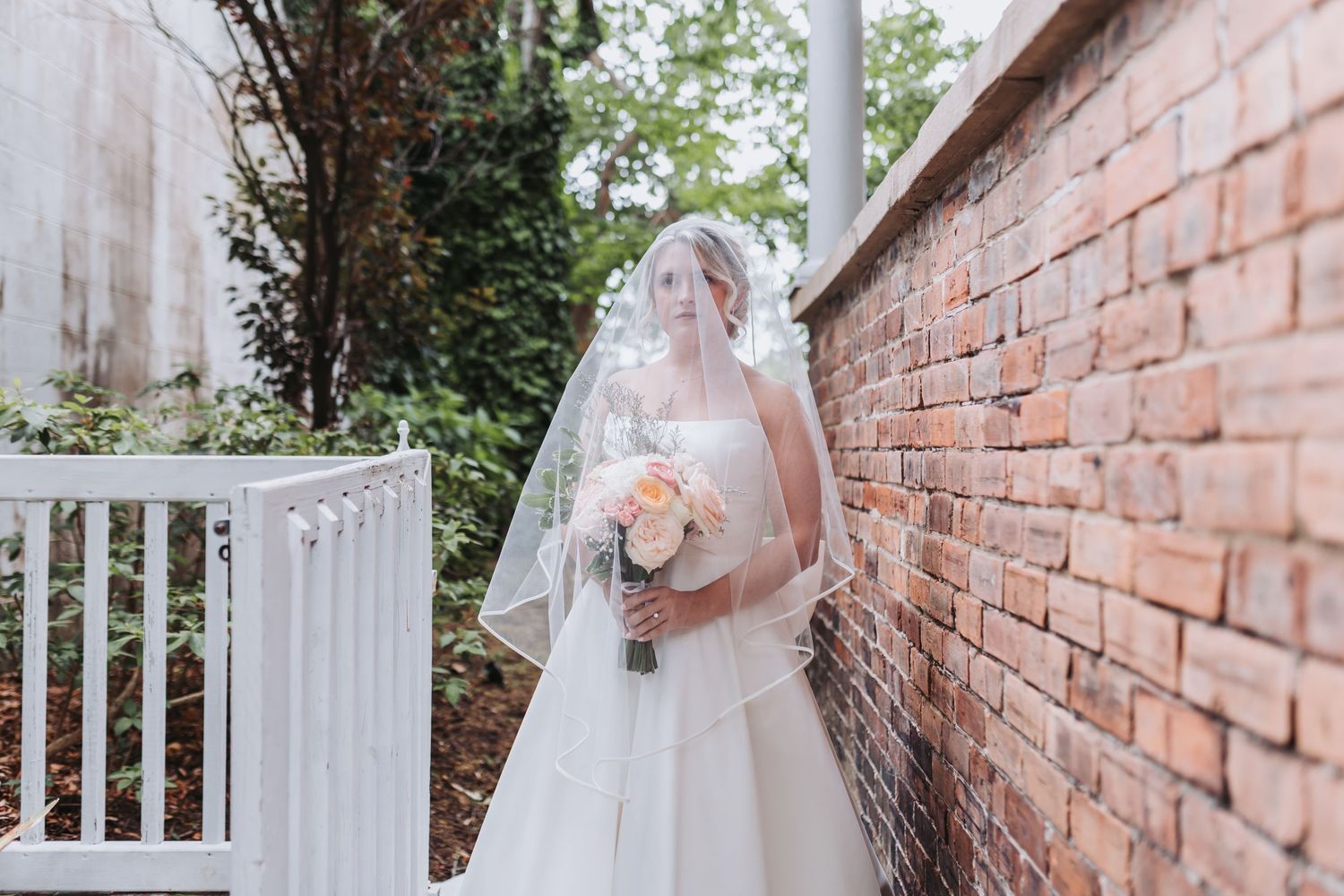 Bride in white gown and veil holding bouquet of pink roses standing beside a rustic brick wall outdoors.
