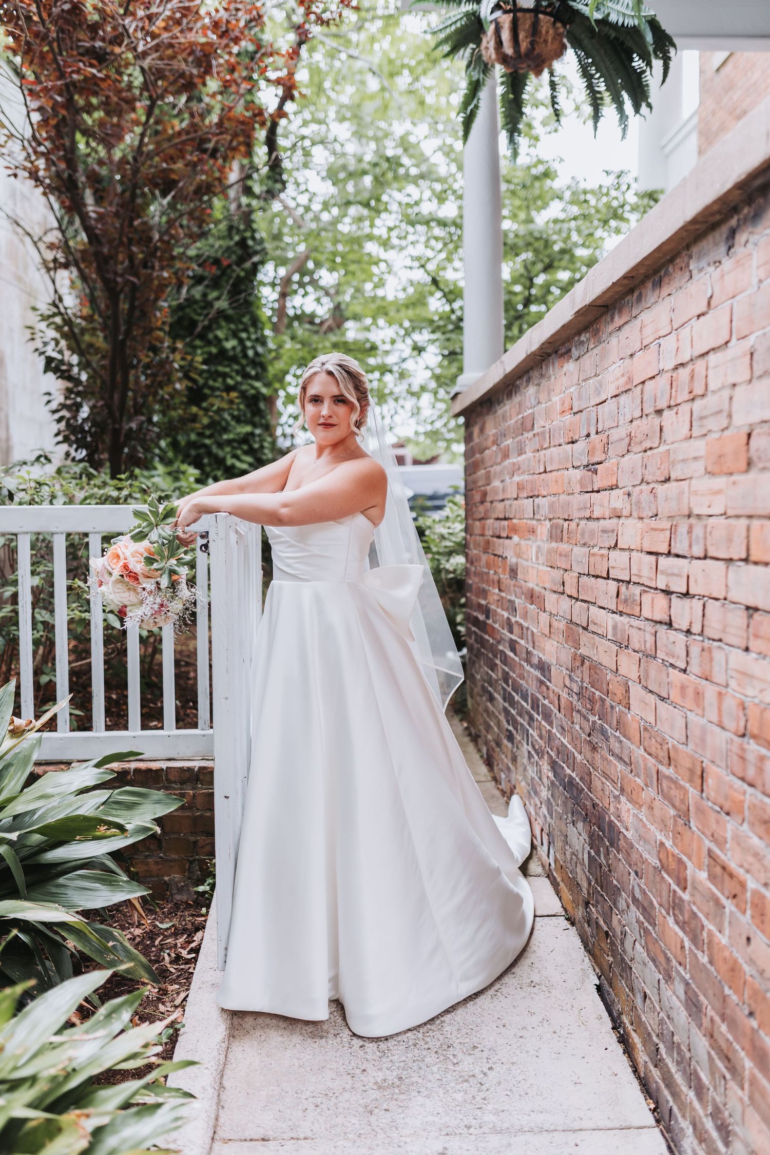 Elegant bride in strapless white gown poses by brick wall with bouquet.