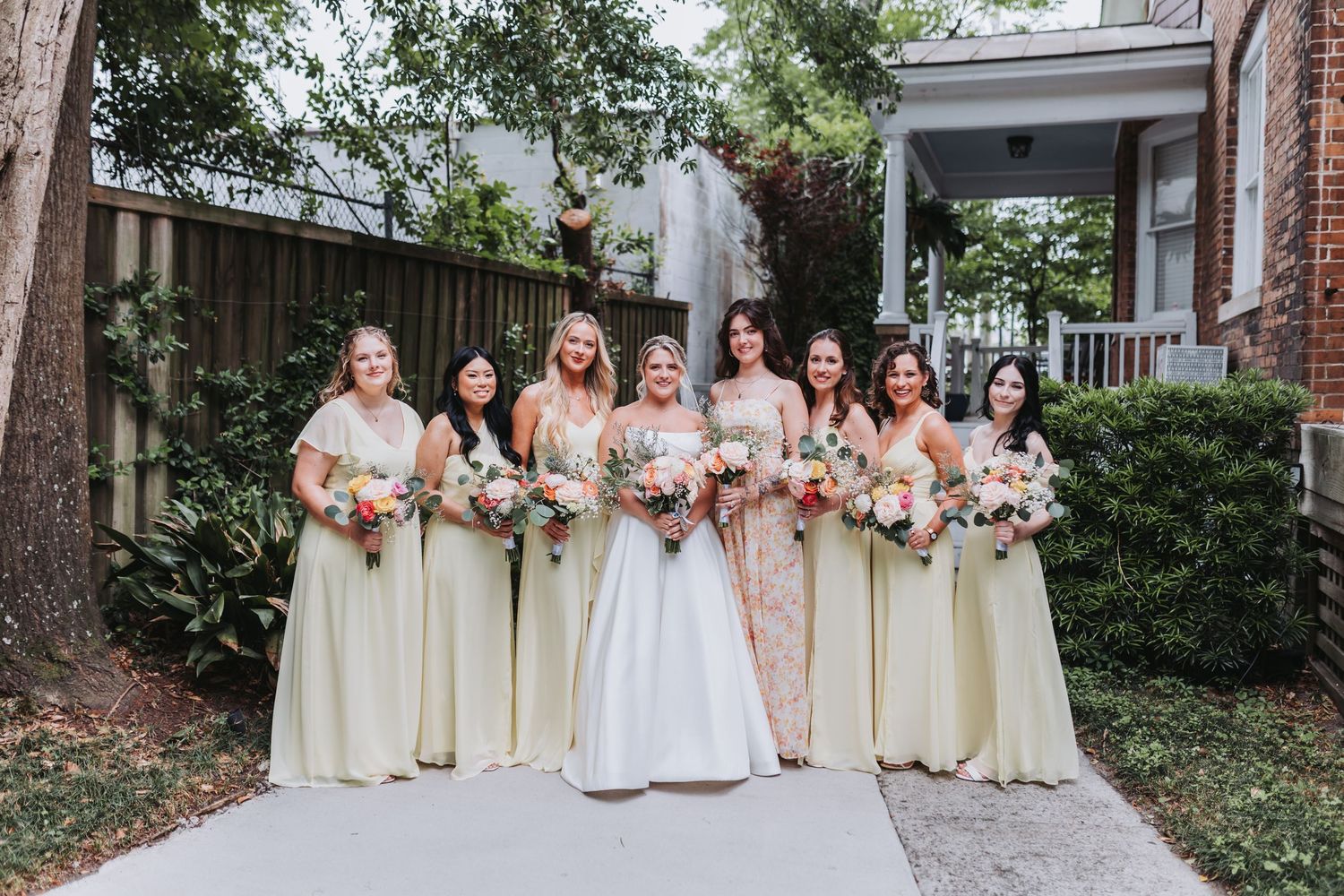 Bride with bridesmaids in yellow dresses holding colorful bouquets outdoors on garden pathway.