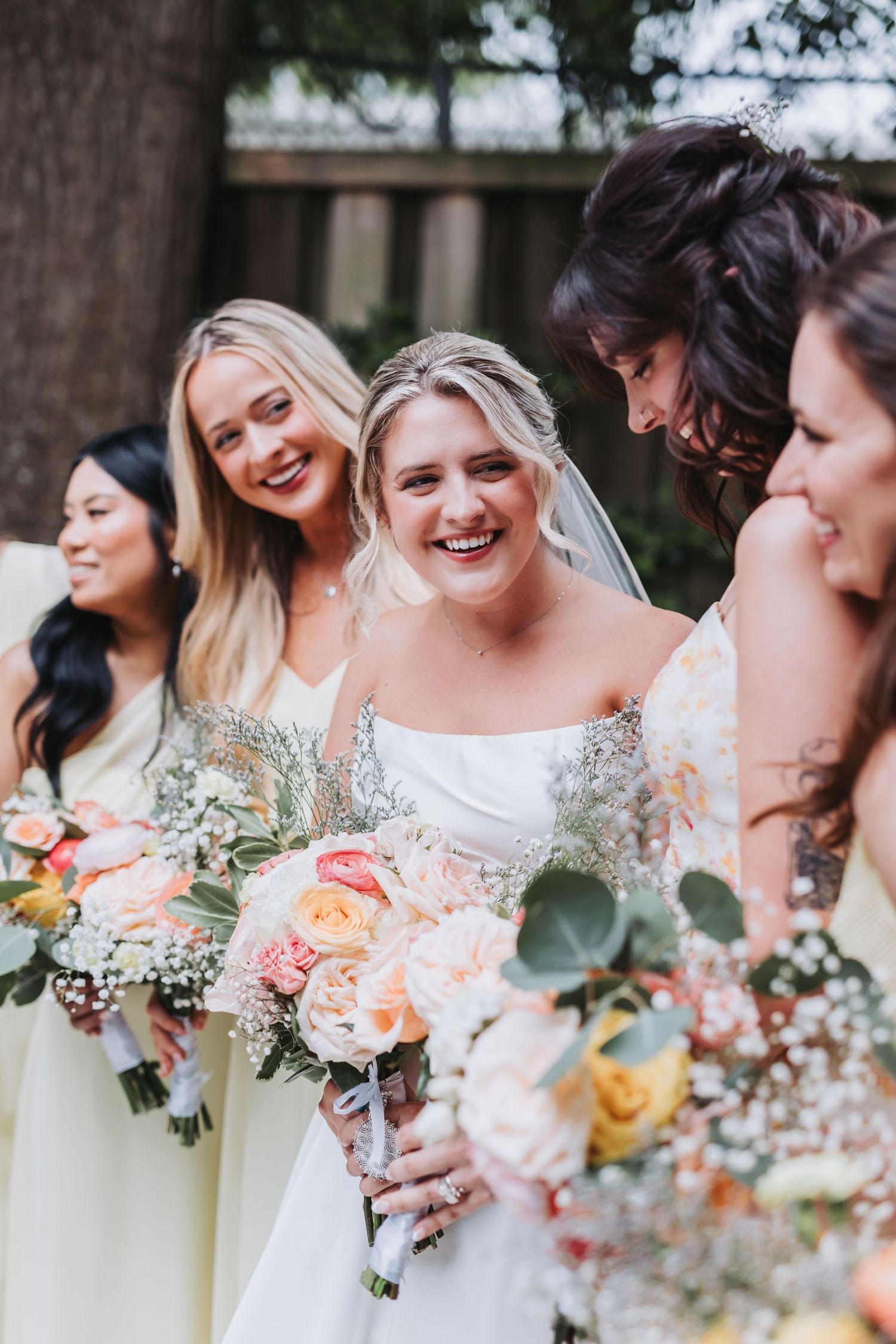 Bride laughing joyfully with bridesmaids in pale yellow dresses holding colorful floral bouquets outdoors.