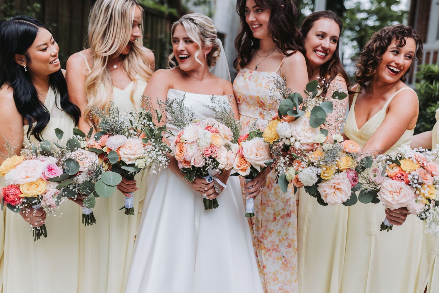 Bride and bridesmaids in yellow dresses laughing together holding pink and peach floral bouquets.