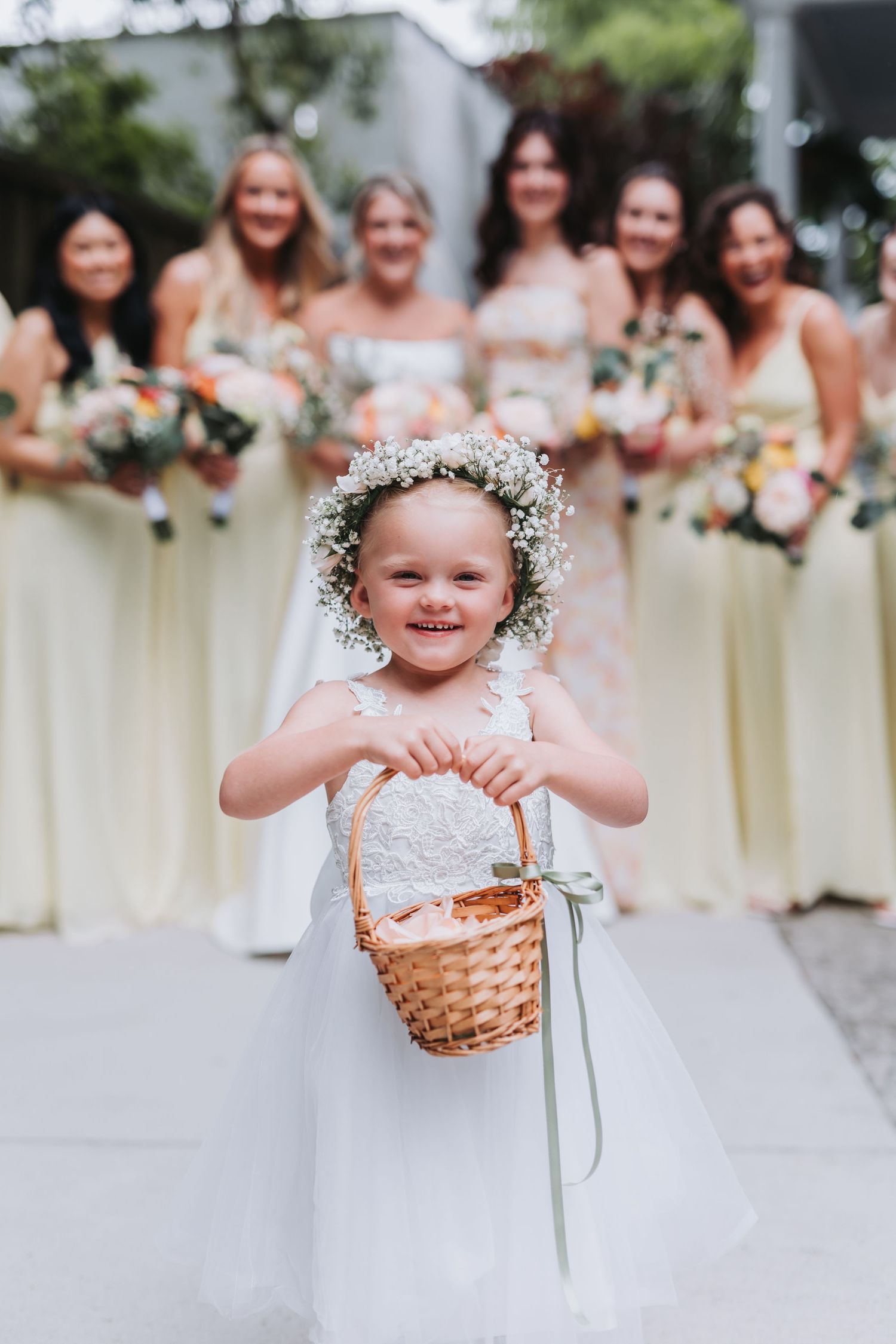 Smiling flower girl in white dress with baby's breath crown holding wicker basket at wedding.