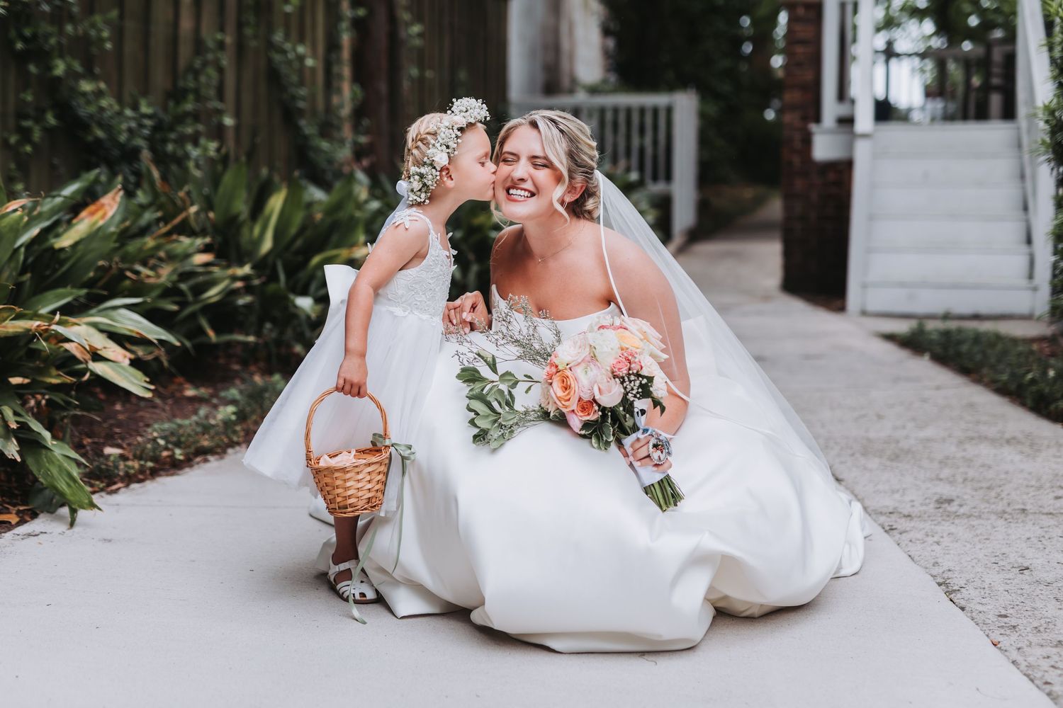 Flower girl kissing smiling bride on cheek as bride kneels on garden pathway holding bouquet.