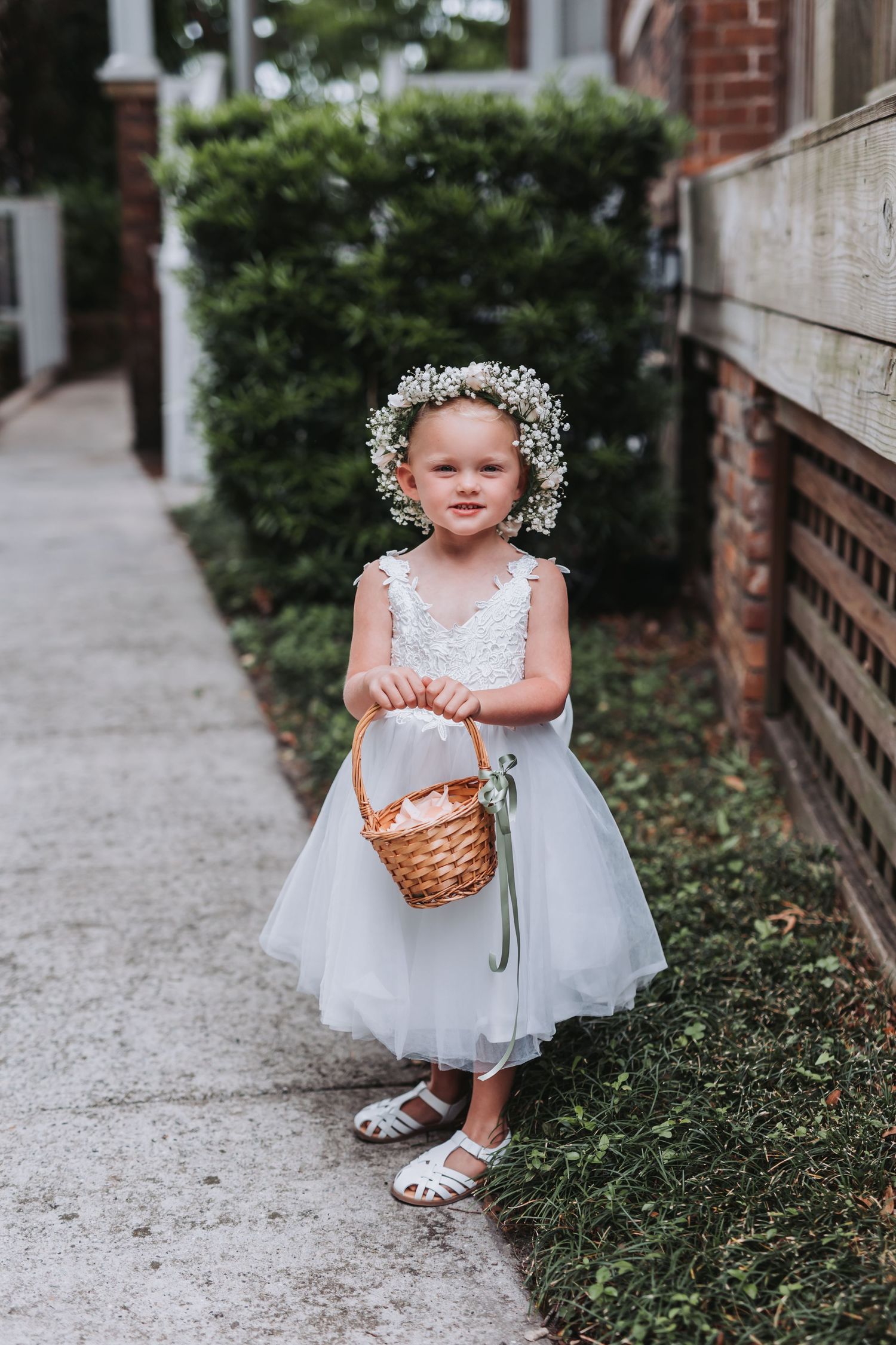 A young flower girl in a white tulle dress and floral crown holds a wicker basket on a garden pathway.
