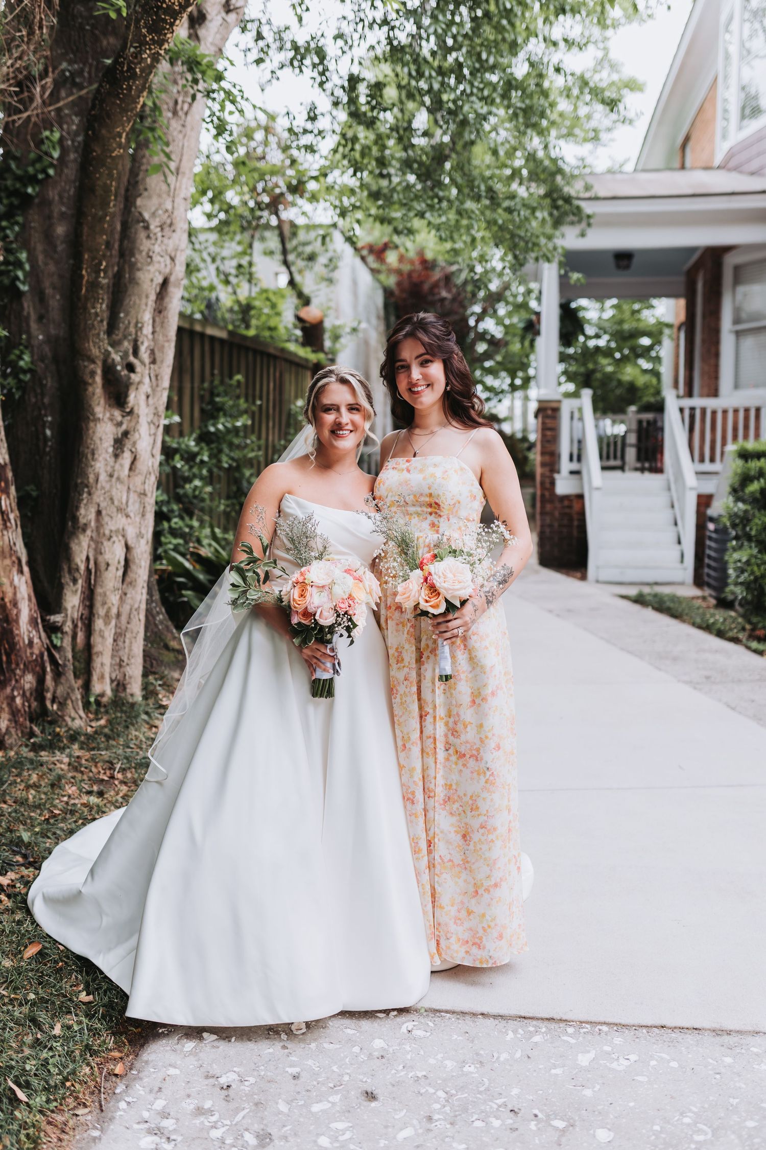 Bride in white ball gown and bridesmaid in floral dress holding bouquets on outdoor pathway.