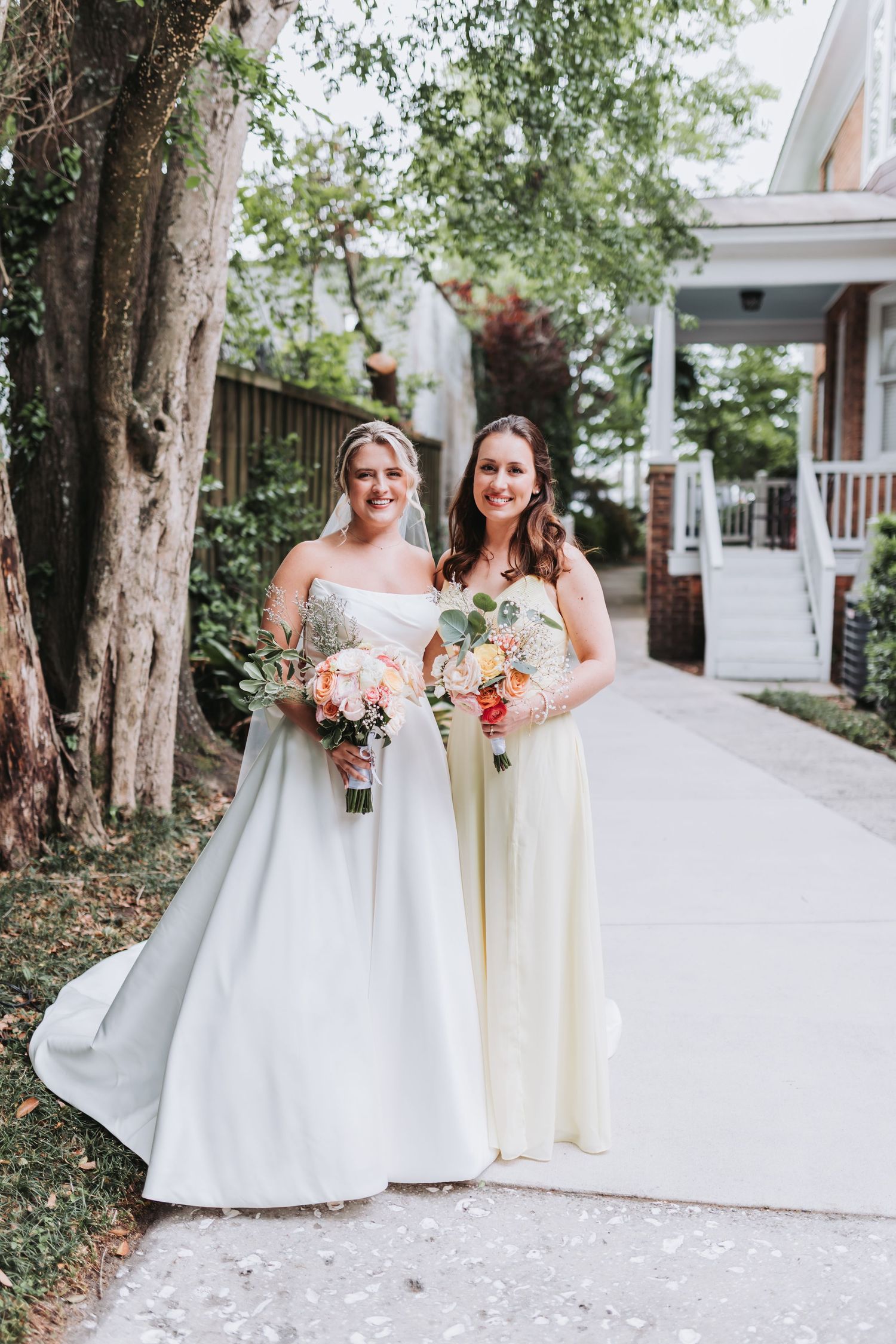 Bride in white gown and bridesmaid in yellow dress holding floral bouquets on a garden pathway.