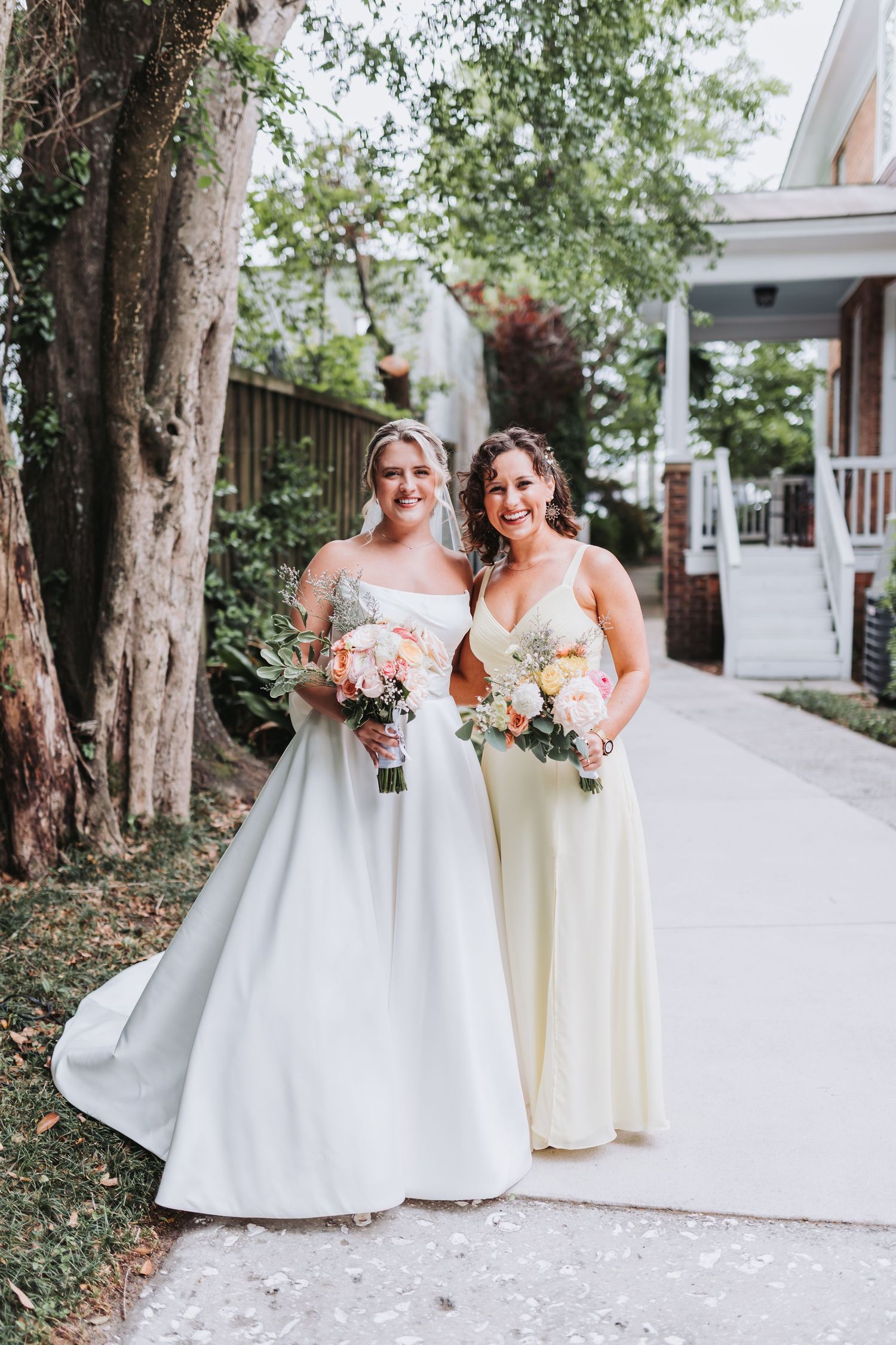 Bride in white gown and bridesmaid in yellow dress holding floral bouquets pose on a garden pathway.
