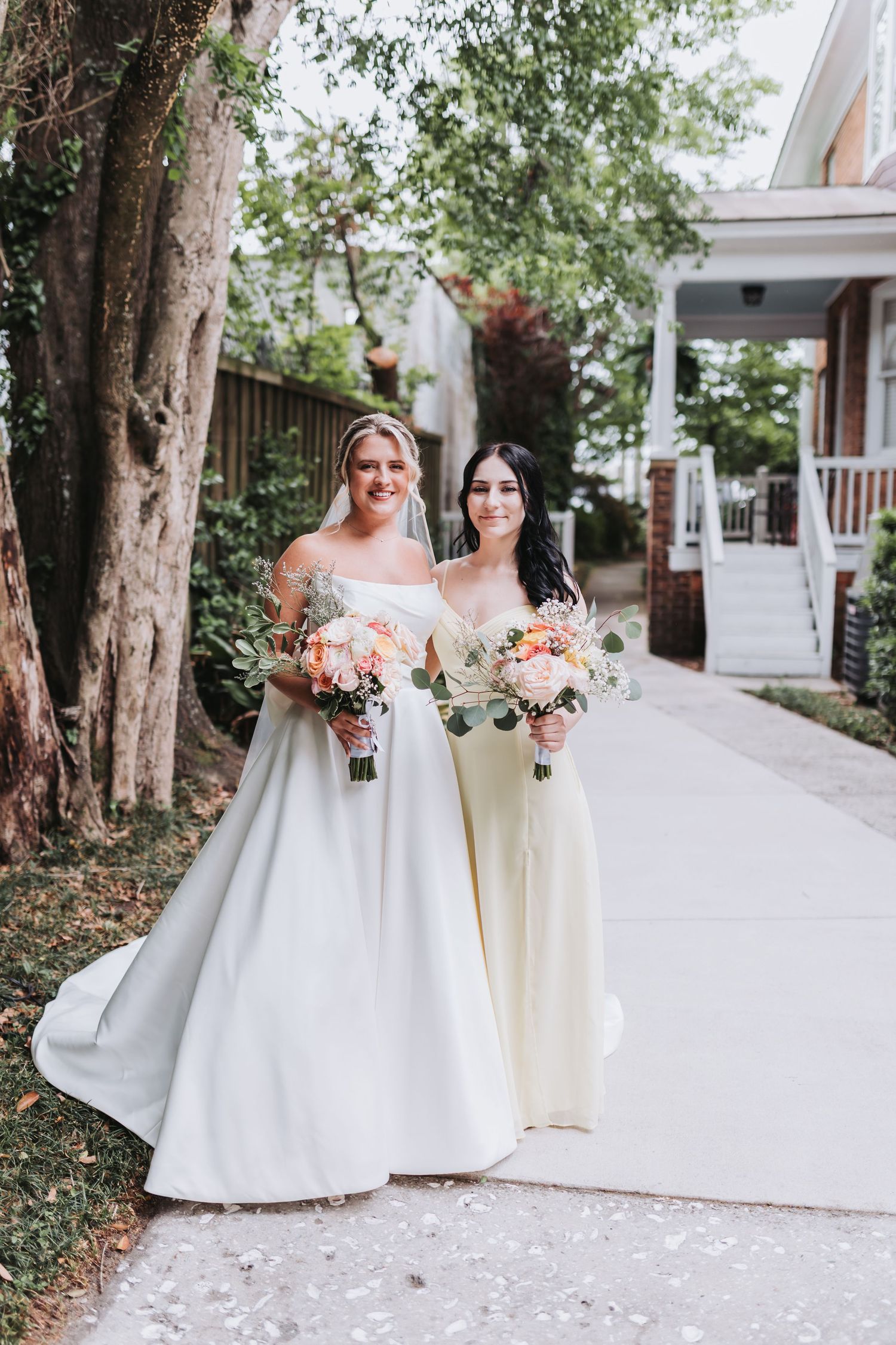 Bride and bridesmaid in pale yellow dress smile together on a tree-lined pathway holding floral bouquets.