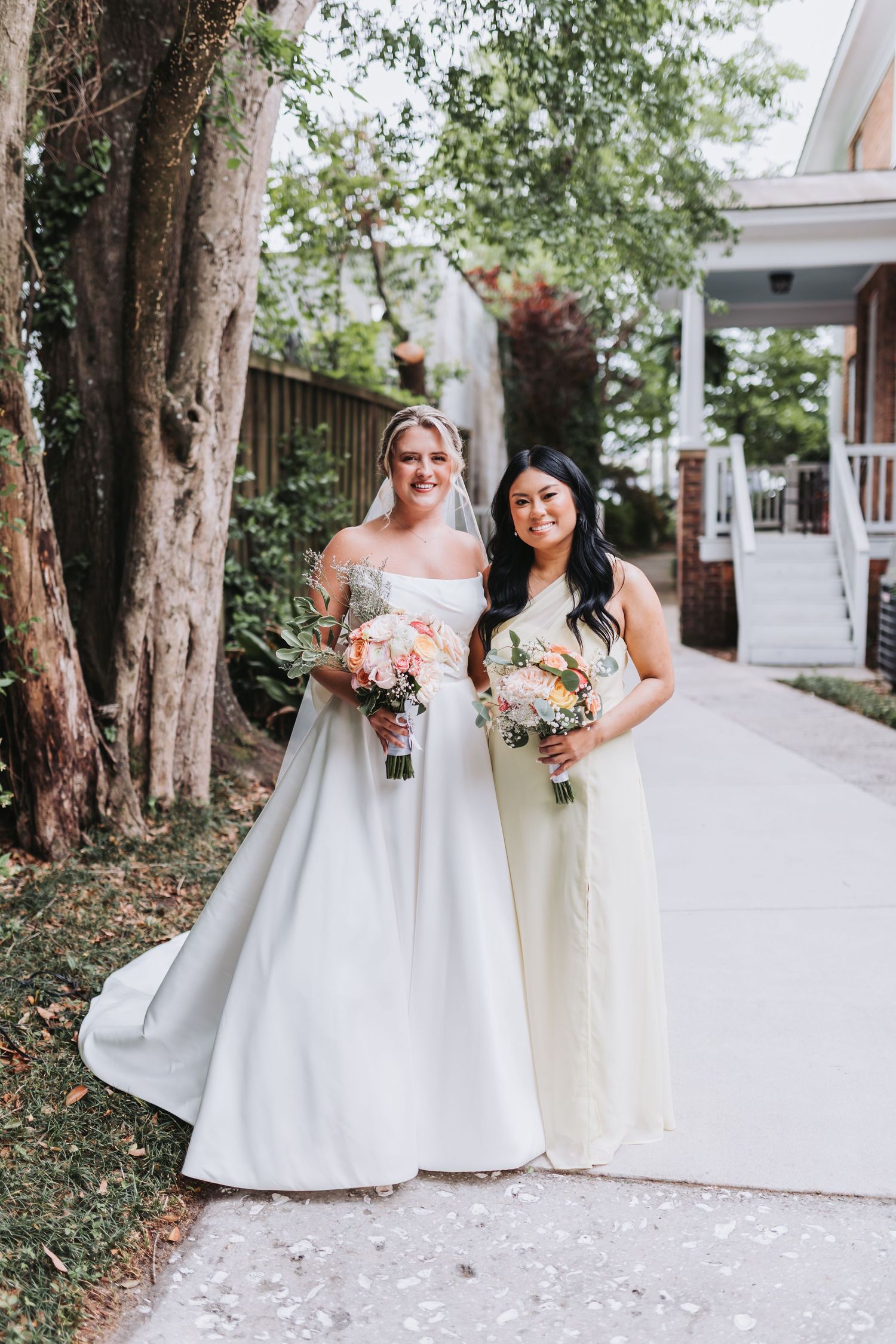 Bride in strapless white gown poses with bridesmaid in yellow dress holding bouquets outdoors.