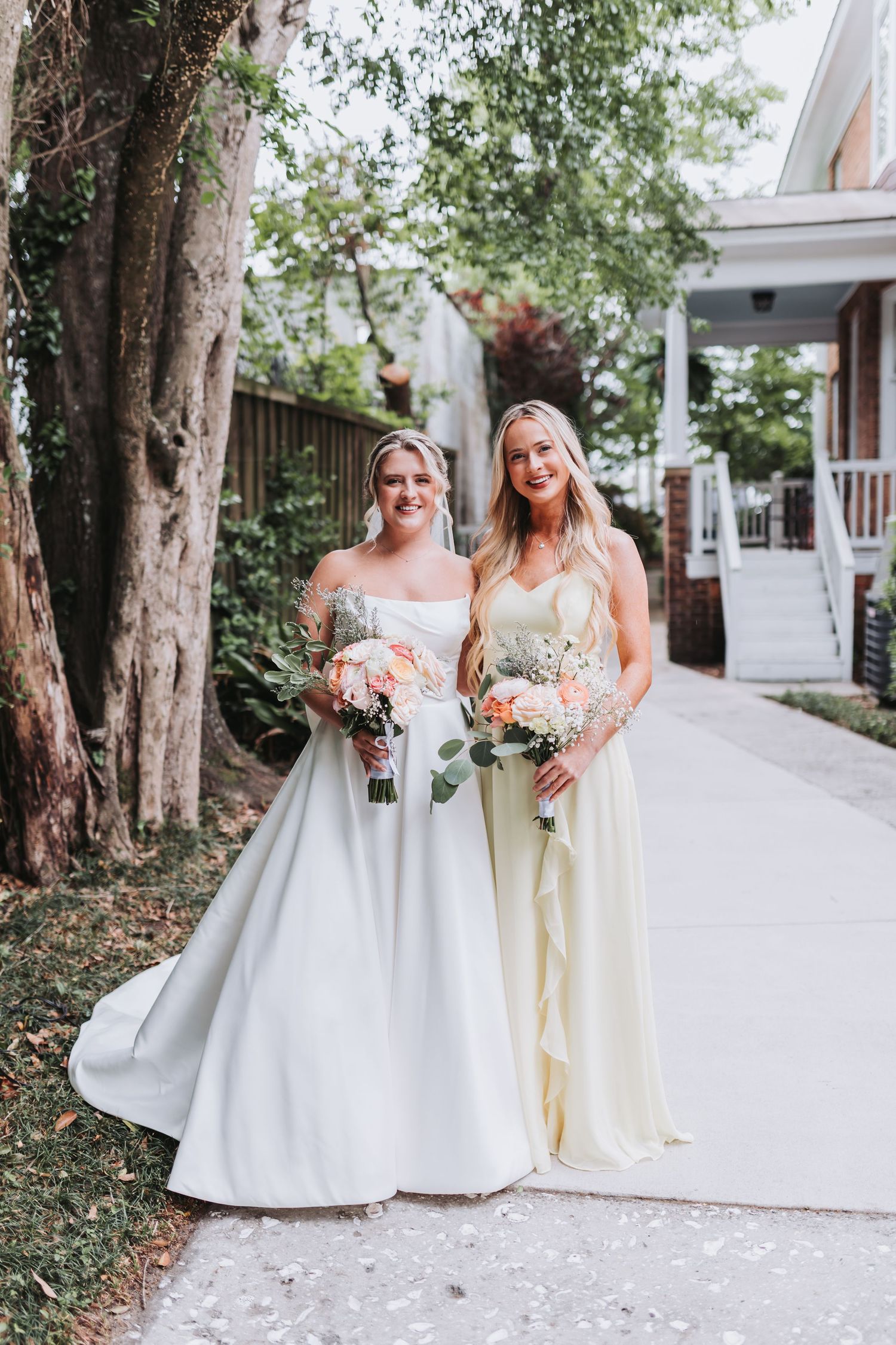 Bride in white ball gown poses with bridesmaid in yellow dress holding bouquets outdoors.