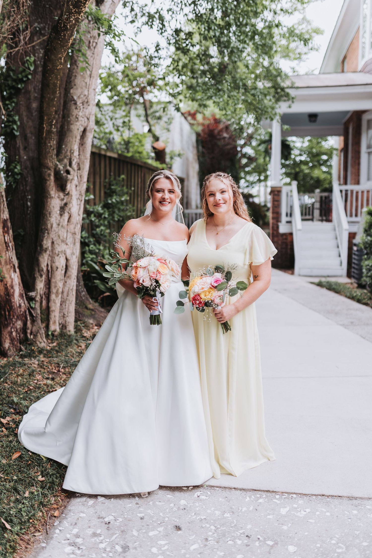 Bride in white strapless ballgown poses with bridesmaid in yellow dress, both holding bouquets outdoors.