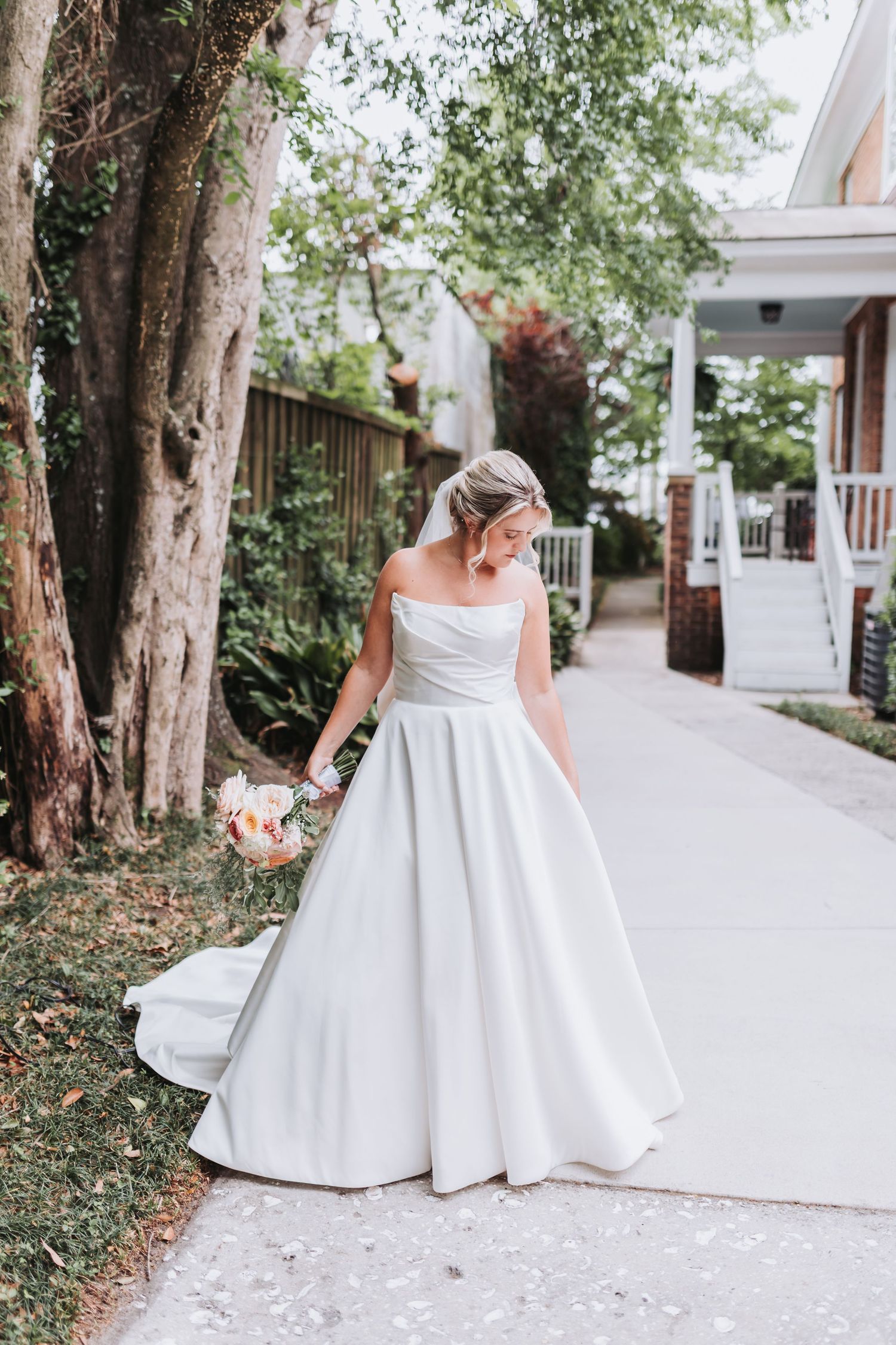 Bride in off-shoulder white ball gown holding bouquet on garden path near trees and white porch.