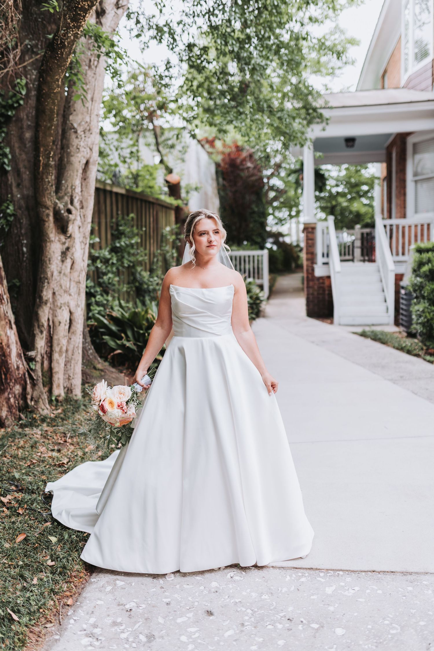 Elegant bride in strapless white ballgown with train holds bouquet on outdoor pathway near trees.