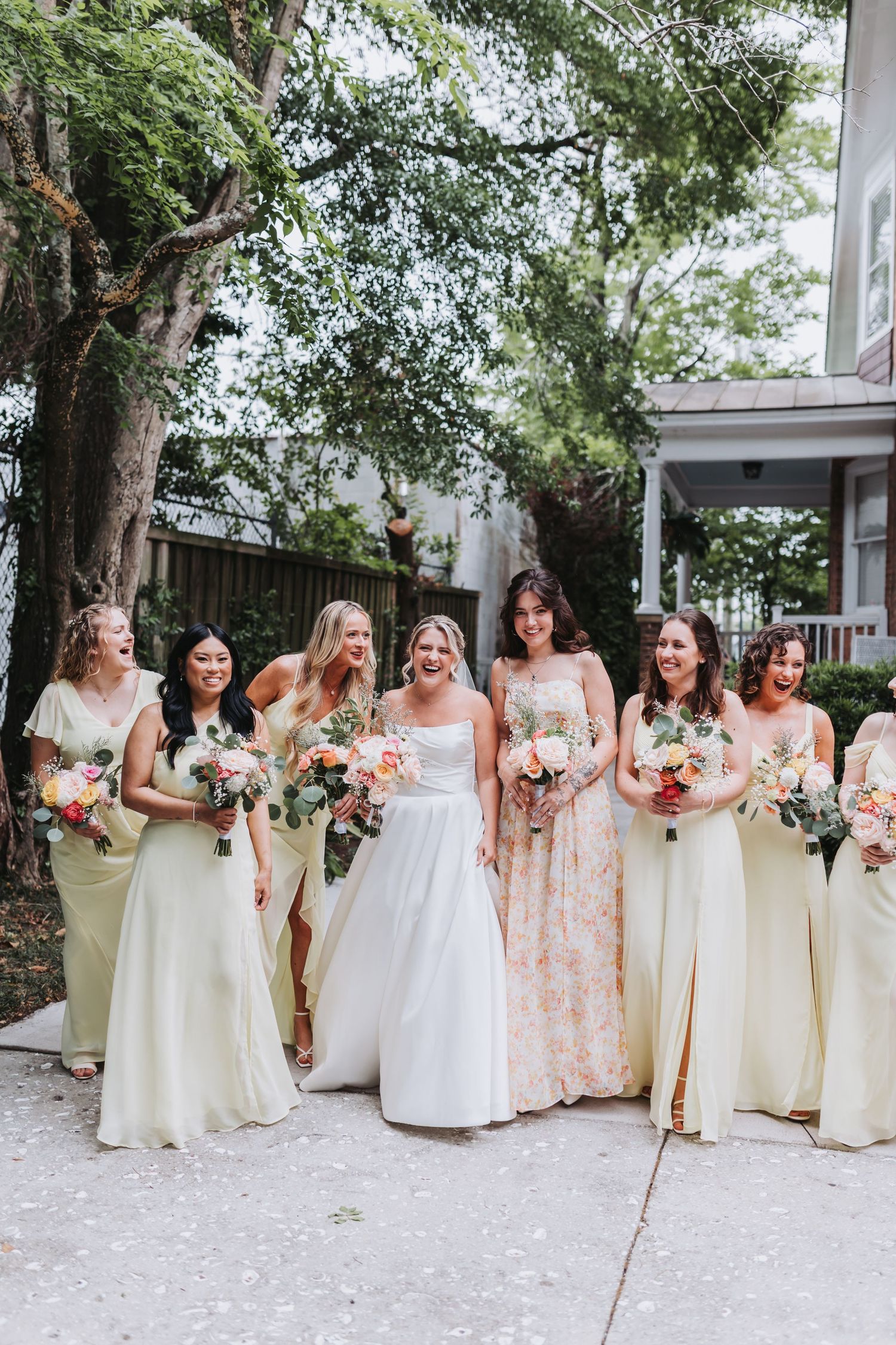 Bride and bridesmaids in pale yellow gowns laughing together outdoors holding colorful floral bouquets.