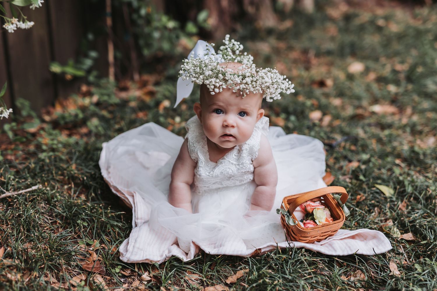 Baby girl in white lace dress and floral crown sits on blanket in garden with Easter basket.