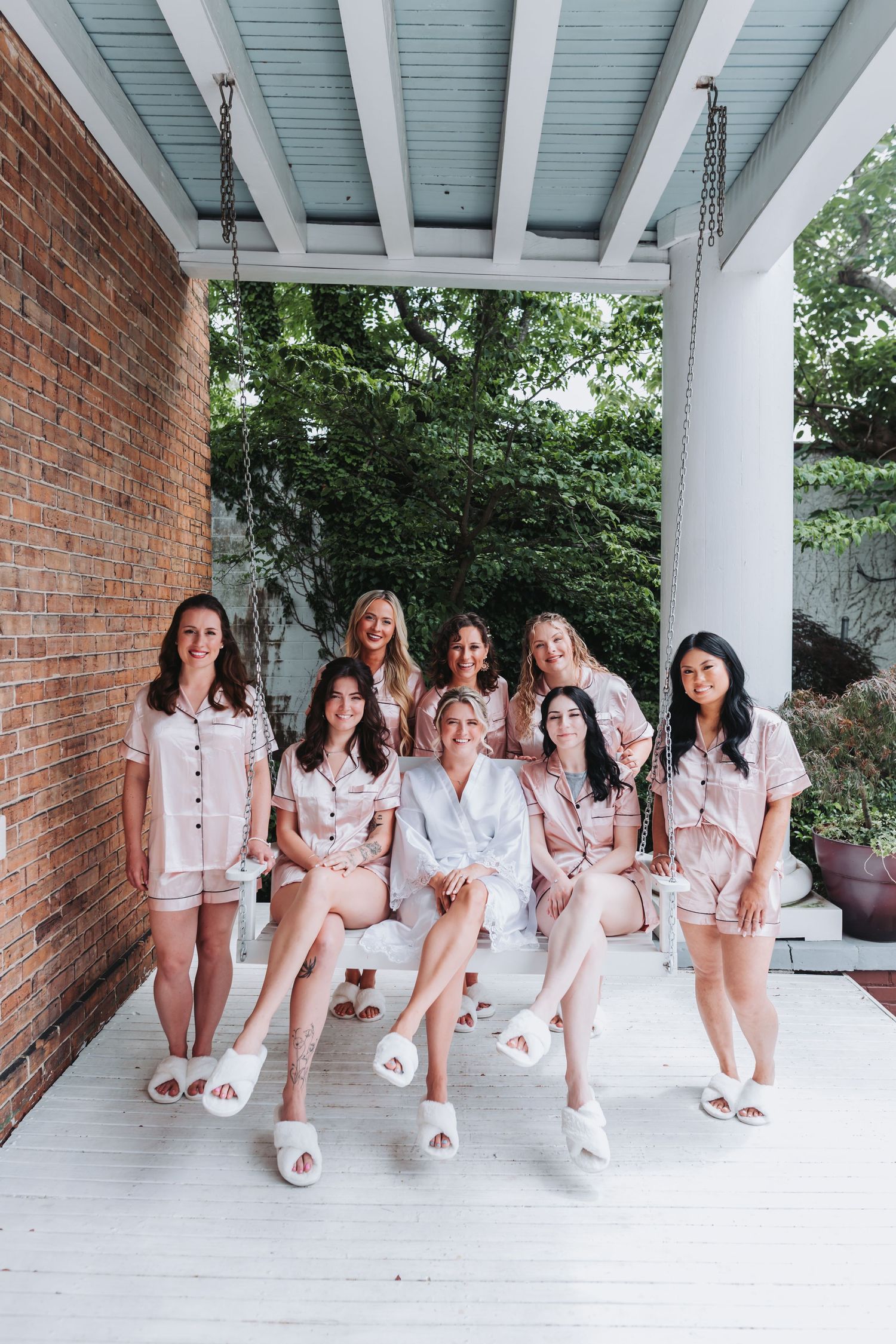 Bridal party in matching pink pajamas and white slippers posing on a porch swing for a wedding morning photo.