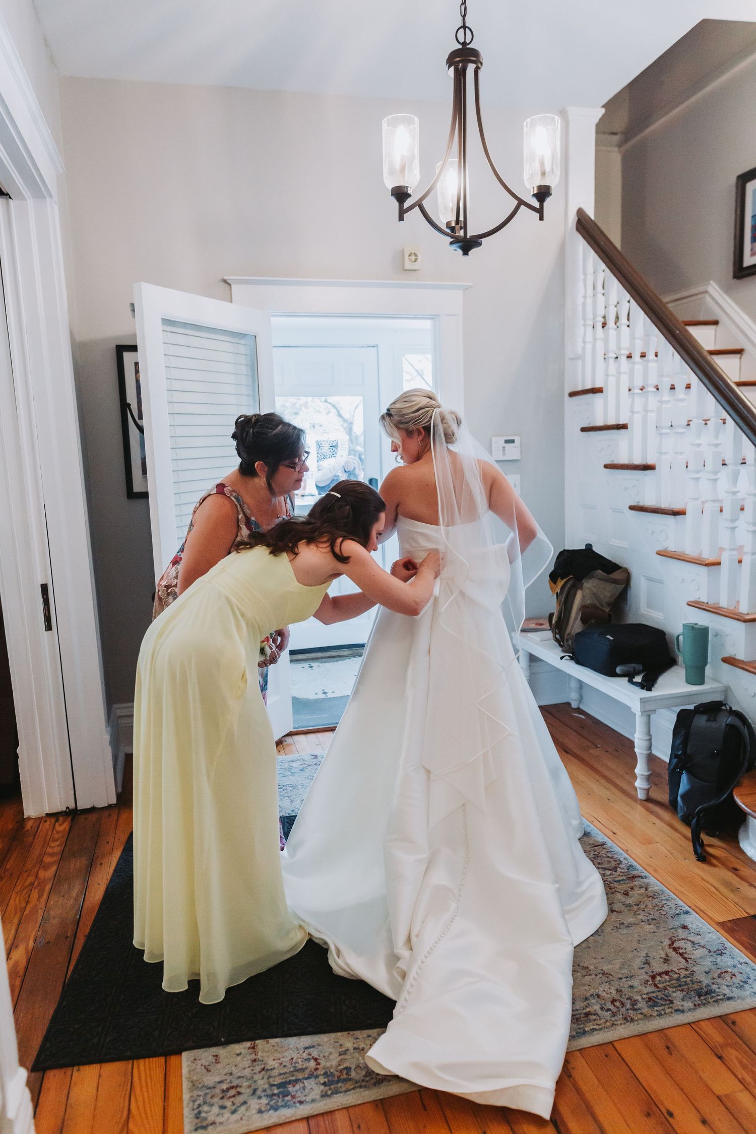 Two bridesmaids in yellow dresses help a bride in a white gown and veil prepare in a home entryway.