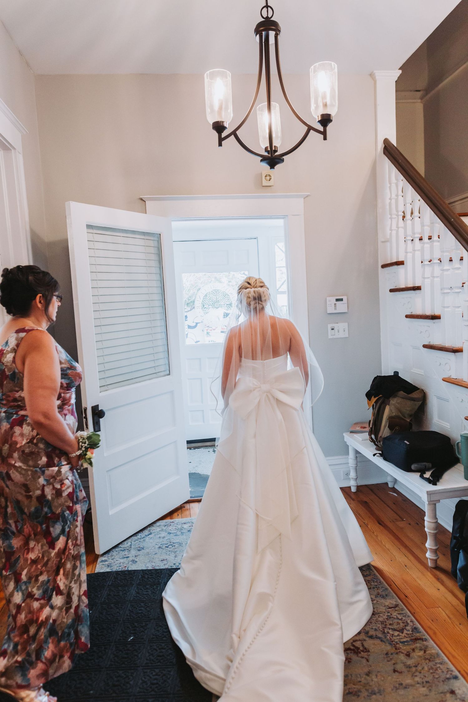Bride in white gown with bow stands at front door as bridesmaid looks on in cozy home entryway.