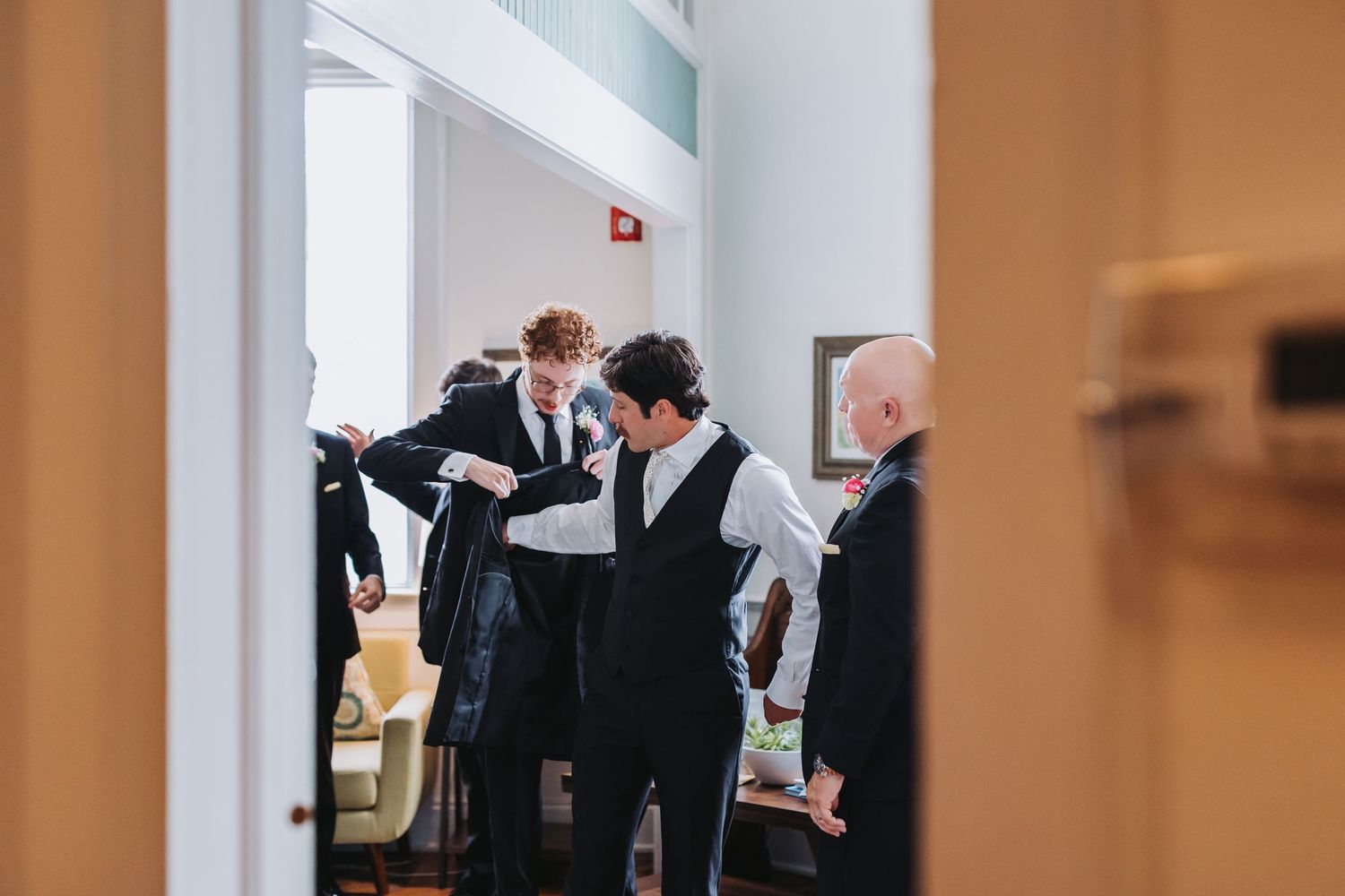 Groomsmen help each other dress in formal black suits before a wedding ceremony indoors.