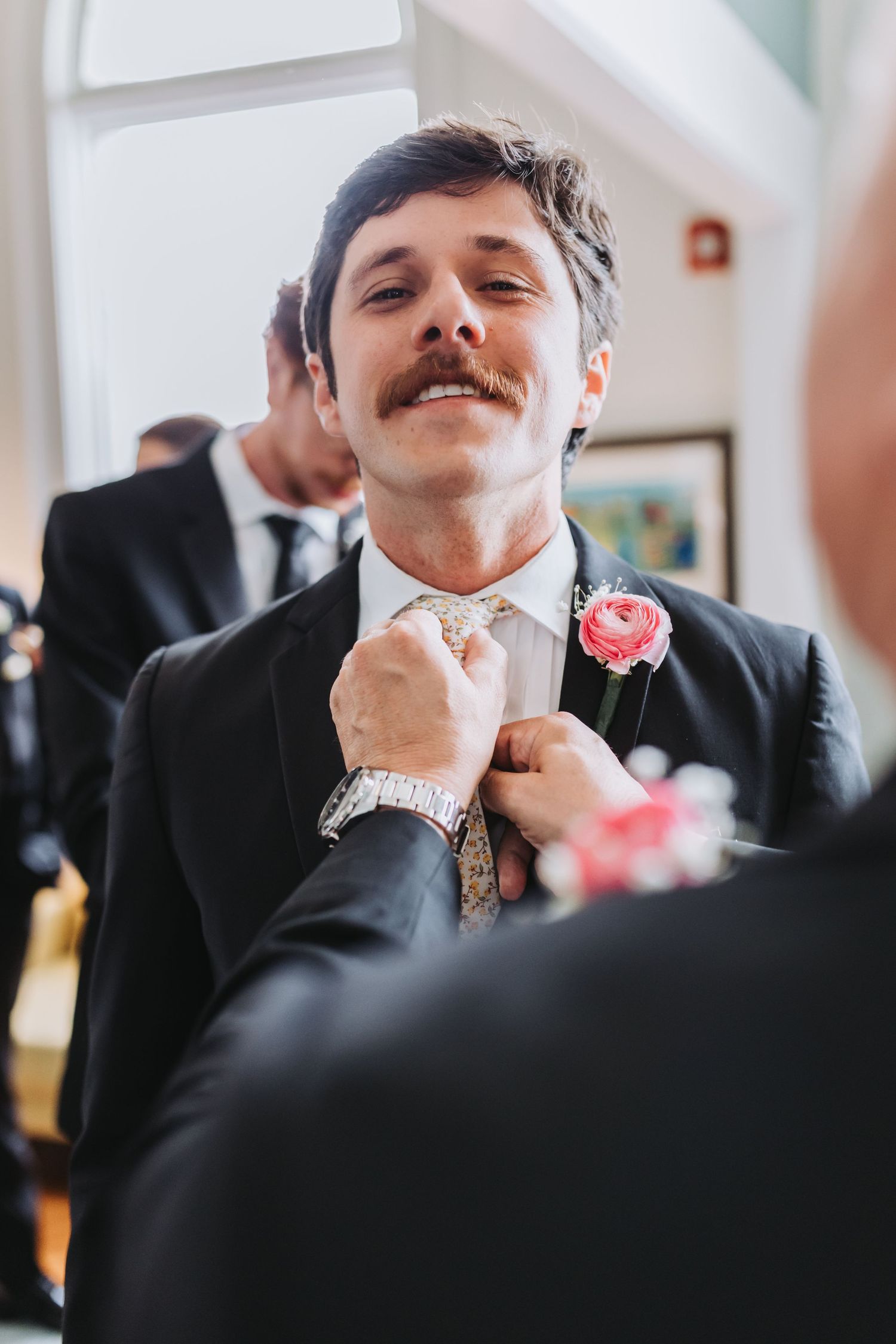 Groom with mustache smiles as boutonniere is pinned to black suit lapel before wedding ceremony.