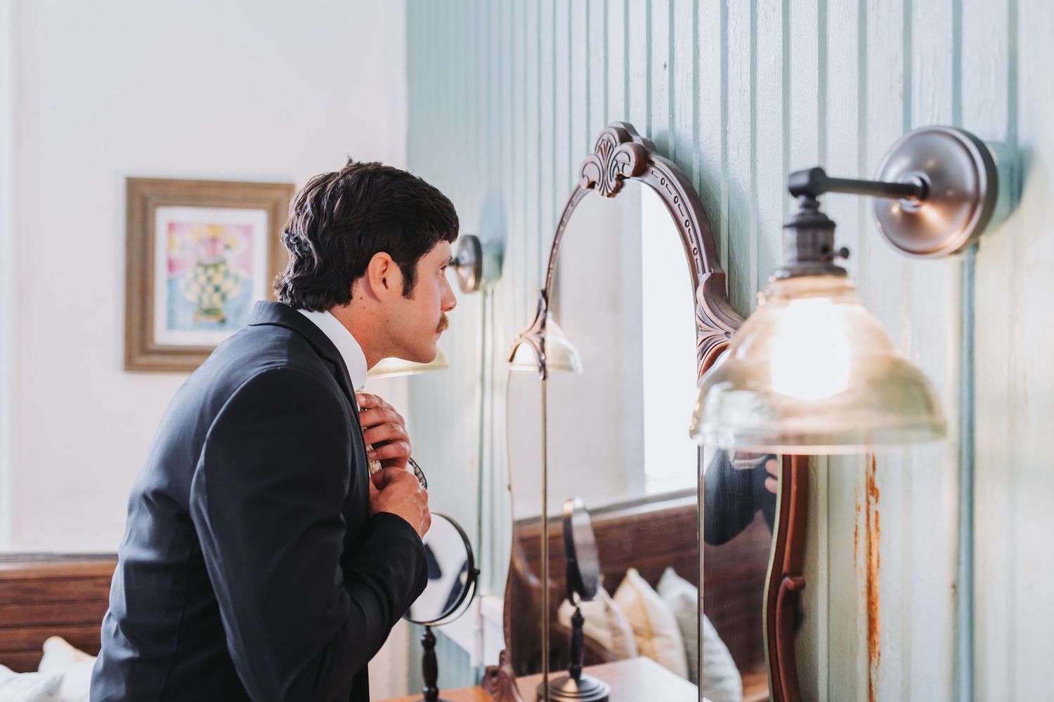 Groom in black suit adjusts tie while looking in mirror during wedding day preparations.
