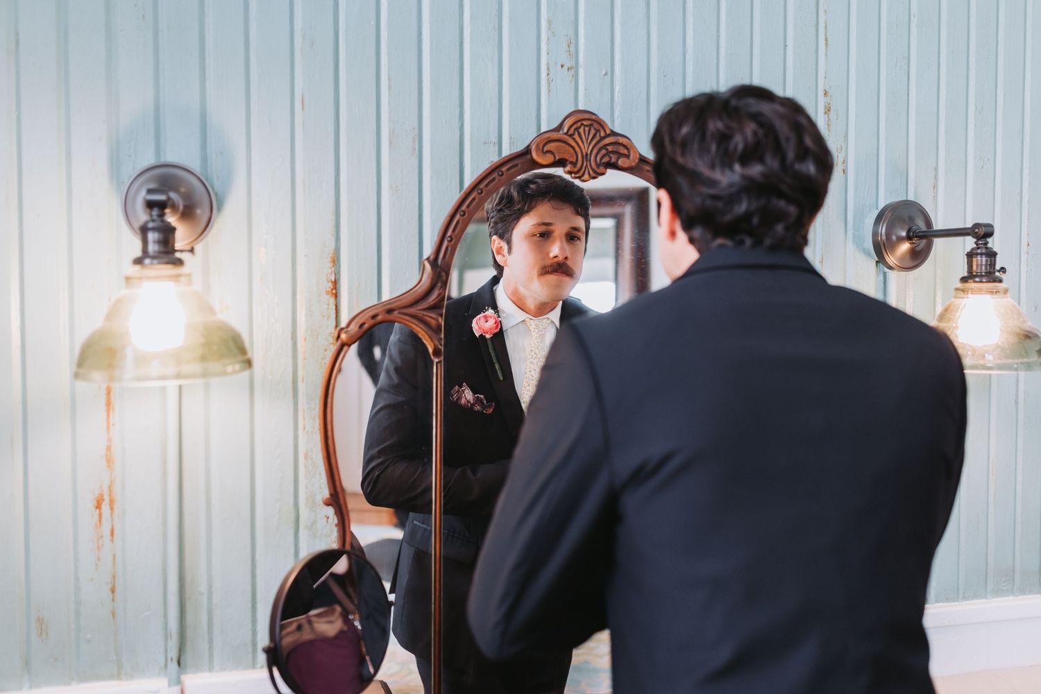 Dapper groom with mustache in dark suit examines his reflection in ornate vintage mirror before wedding.